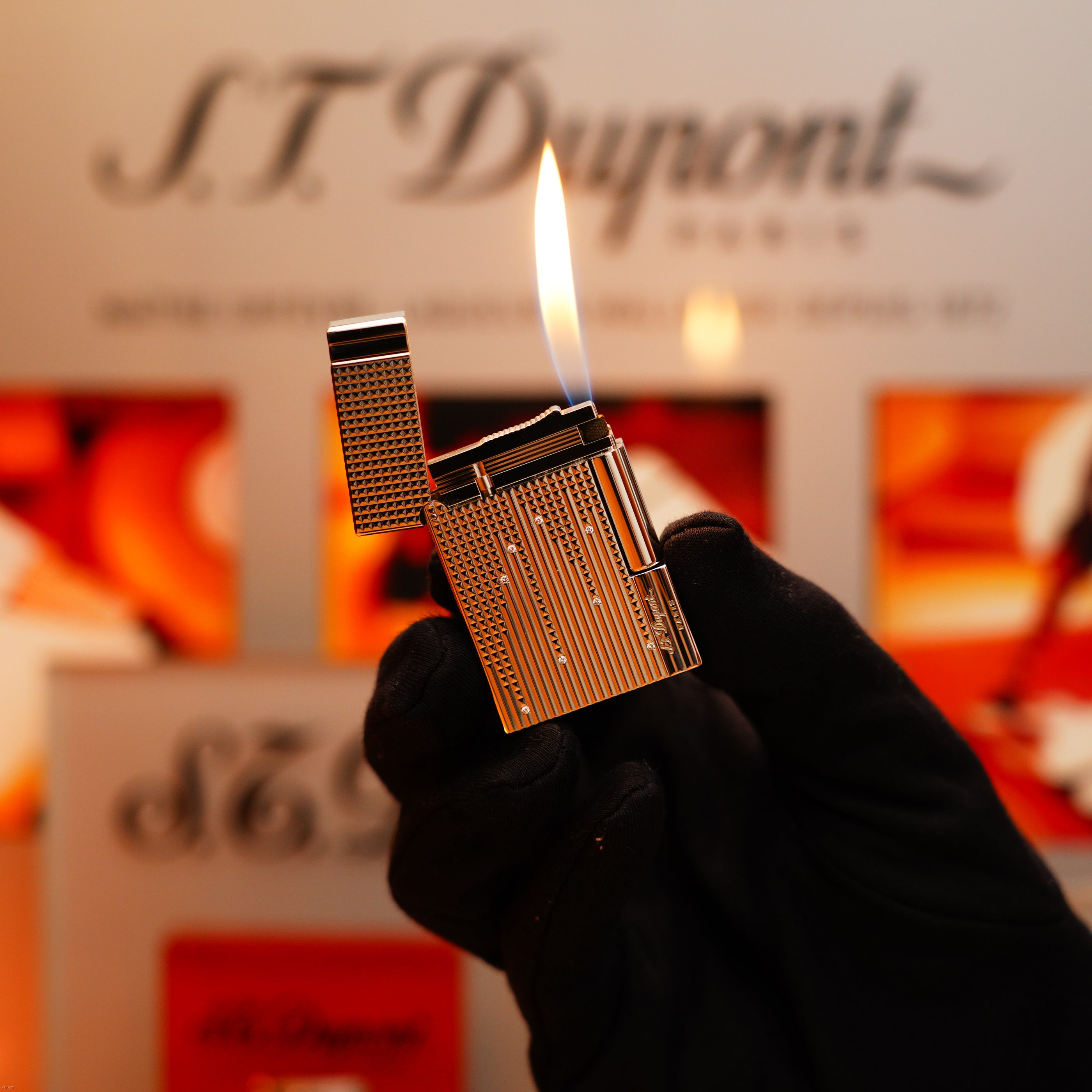 A gloved hand holds a lit Vintage 2005 S.T. Dupont Gatsby Diamond Drops with 16 Natural Diamond Heavy Platinum Limited Edition Lighter before a blurred S.T. Dupont sign and colorful background.