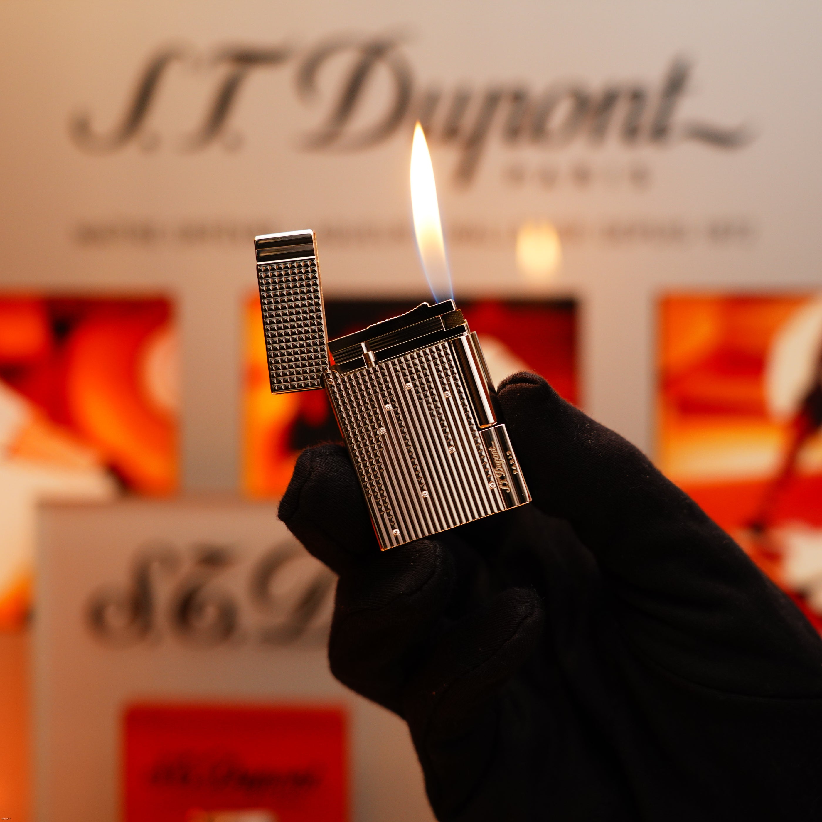 A gloved hand holds an open Vintage 2005 S.T. Dupont Gatsby Diamond Drops with 16 Natural Diamonds Heavy Platinum Limited Edition Lighter, its flame visible against a blurred S.T. Dupont branded background.