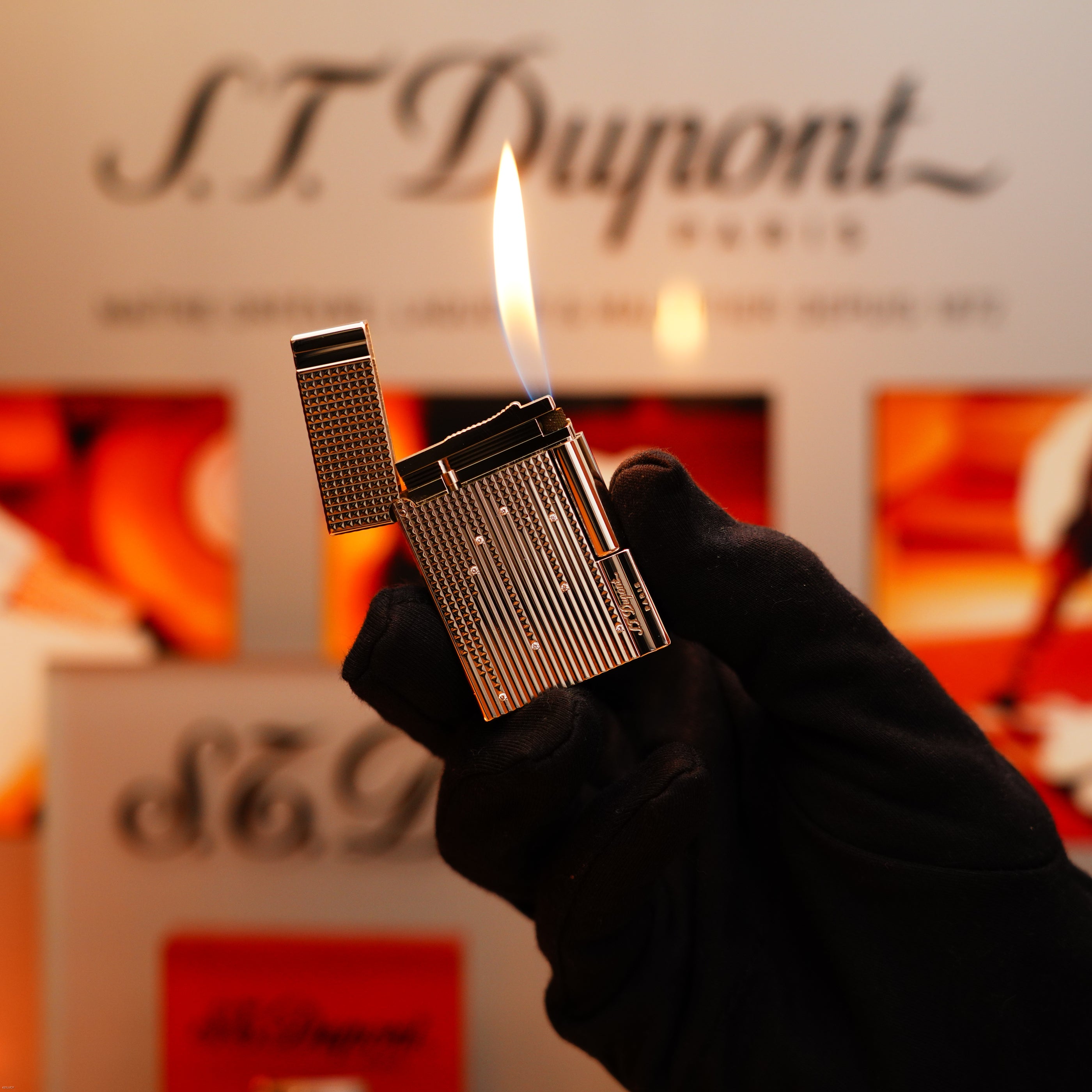 A gloved hand holds a lit S.T. Dupont Vintage 2005 Gatsby Diamond Drops platinum lighter with 16 natural diamonds, in front of a blurred S.T. Dupont Paris sign.