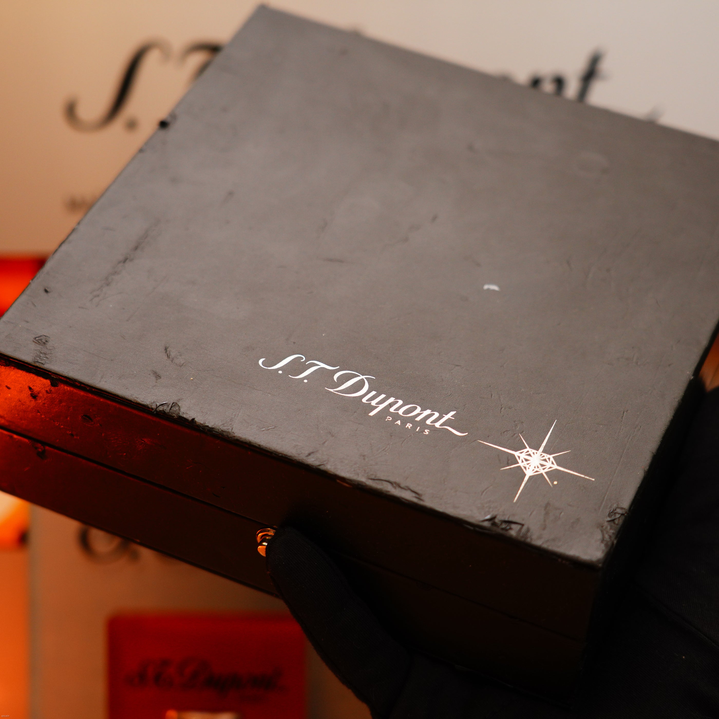 A gloved hand holds a black box with the S.T. Dupont logo, set against a blurred branded background, showcasing the Vintage 2005 S.T. Dupont Gatsby Diamond Drops with 16 Natural Diamond Heavy Platinum Limited Edition Lighter.
