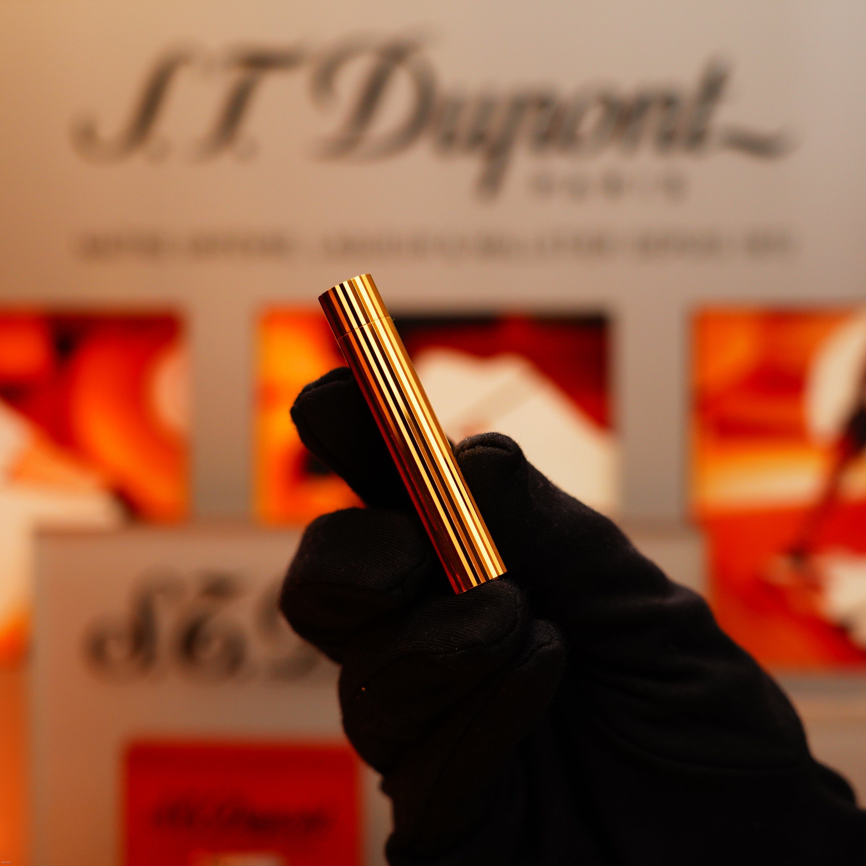 A gloved hand holds a Cartier Vintage 1980 Oval Le Must Lighter with a heavy 24k gold vertical line finish upright, with a blurred sign and packaging in the background.