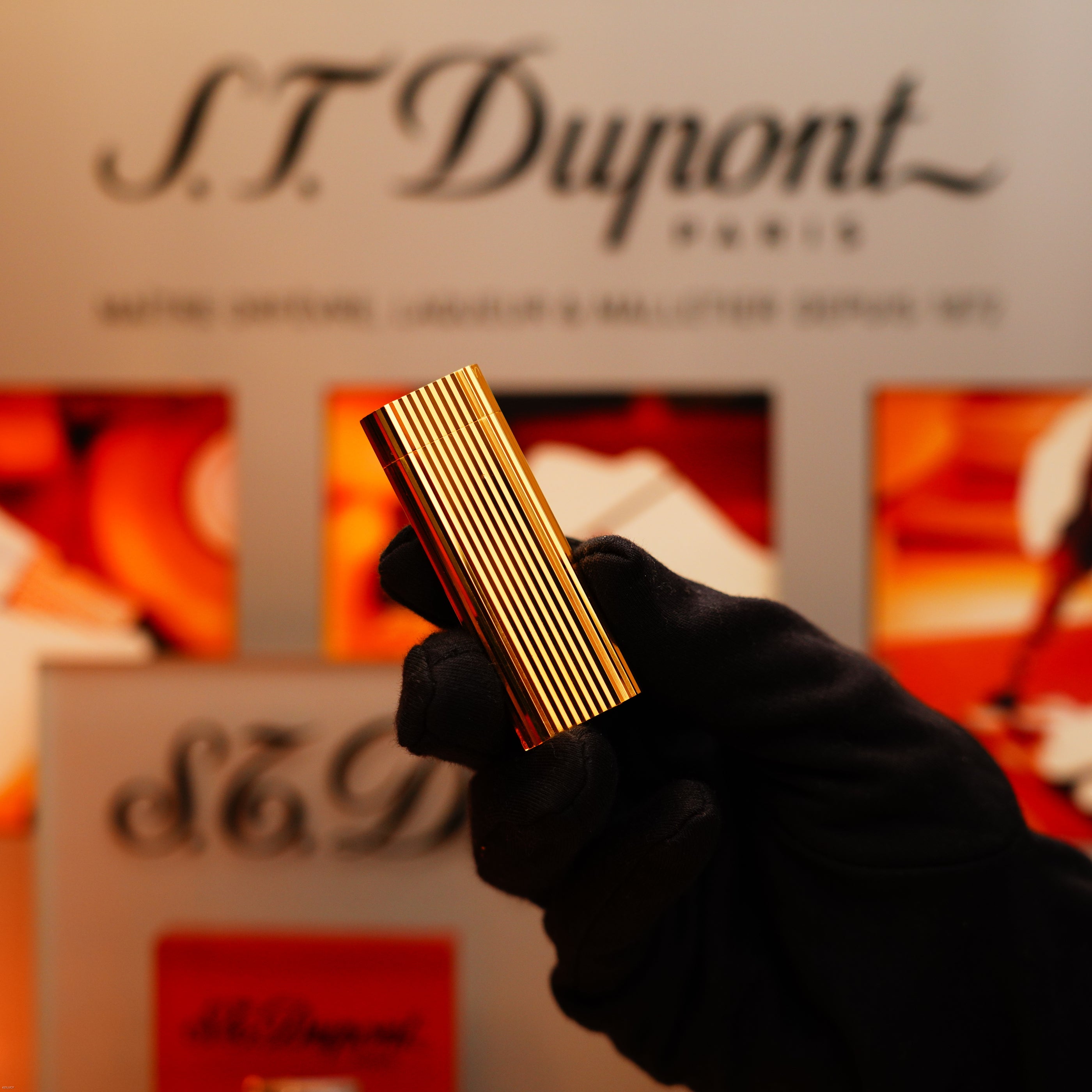 A gloved hand holds a Vintage 1980 Cartier Vertical Line Heavy 24k Gold Finish Oval Le Must Lighter by Cartier, displayed against a warm, orange-toned background.