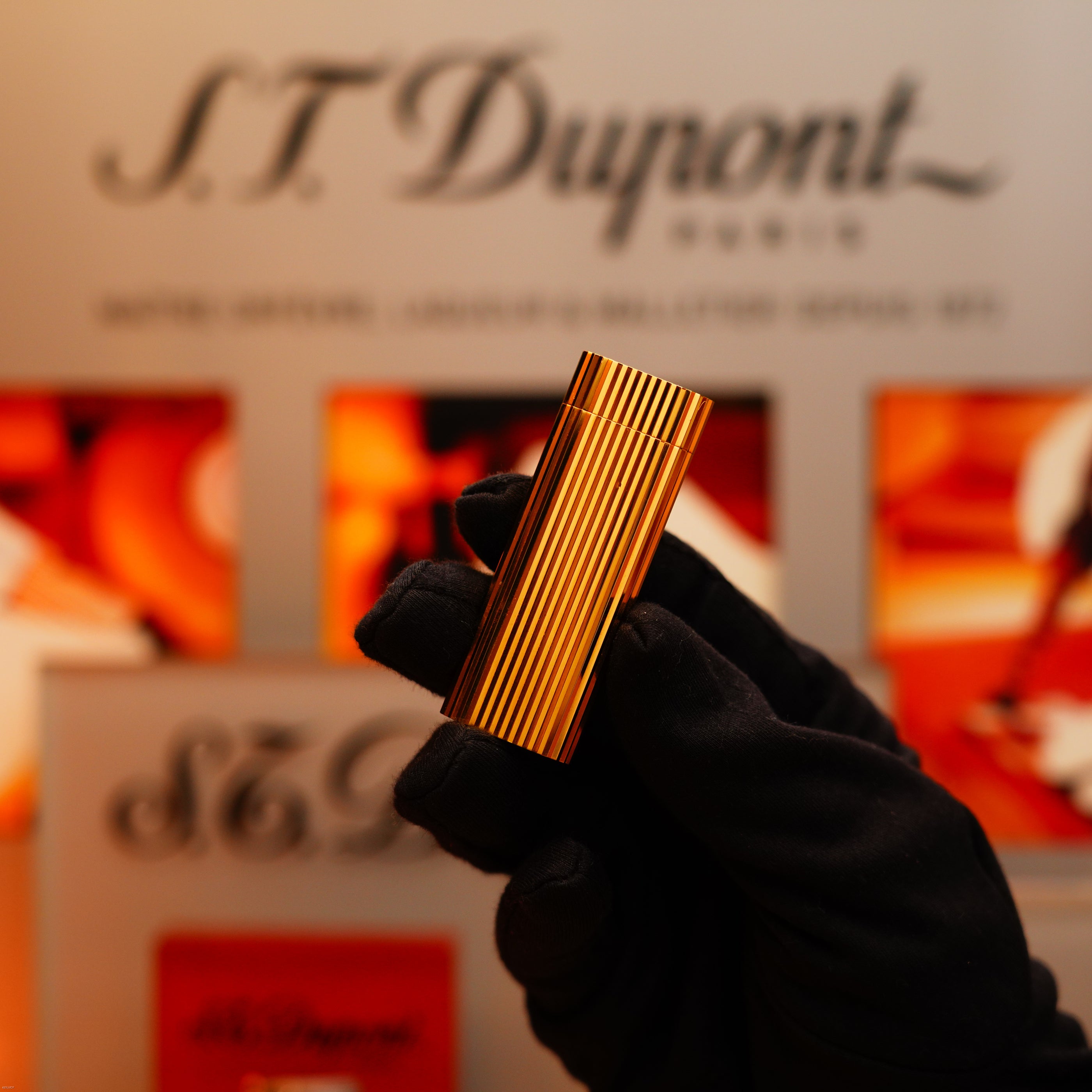 A gloved hand holds a Vintage 1980 Cartier Vertical Line Heavy 24k Gold Finish Oval Le Must Lighter, with a blurred Cartier sign and packaging in the background.