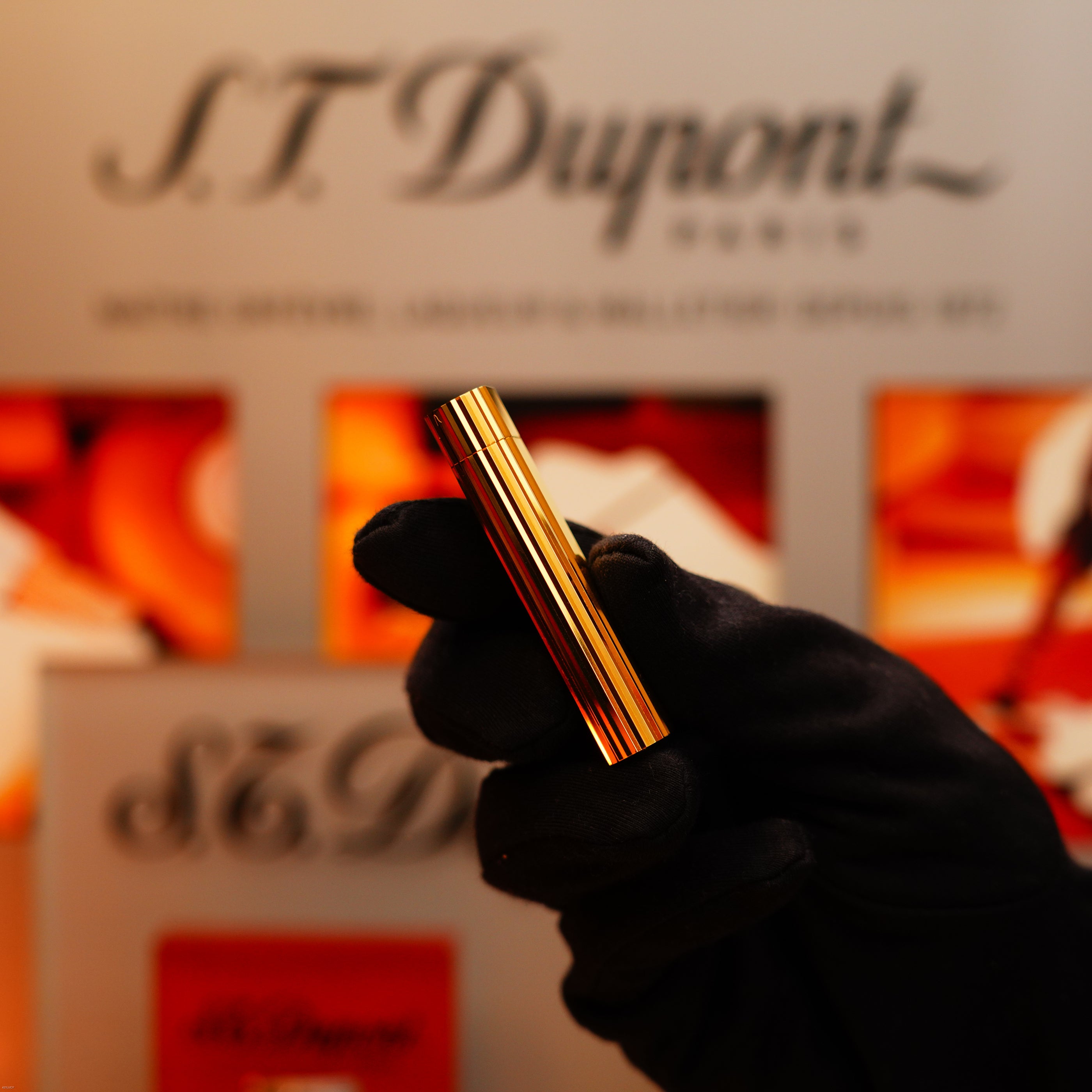 A gloved hand holds the Vintage 1980 Cartier Vertical line Heavy 24k Gold Finish Oval Le Must Lighter, with a blurred Cartier sign and orange background elements behind.