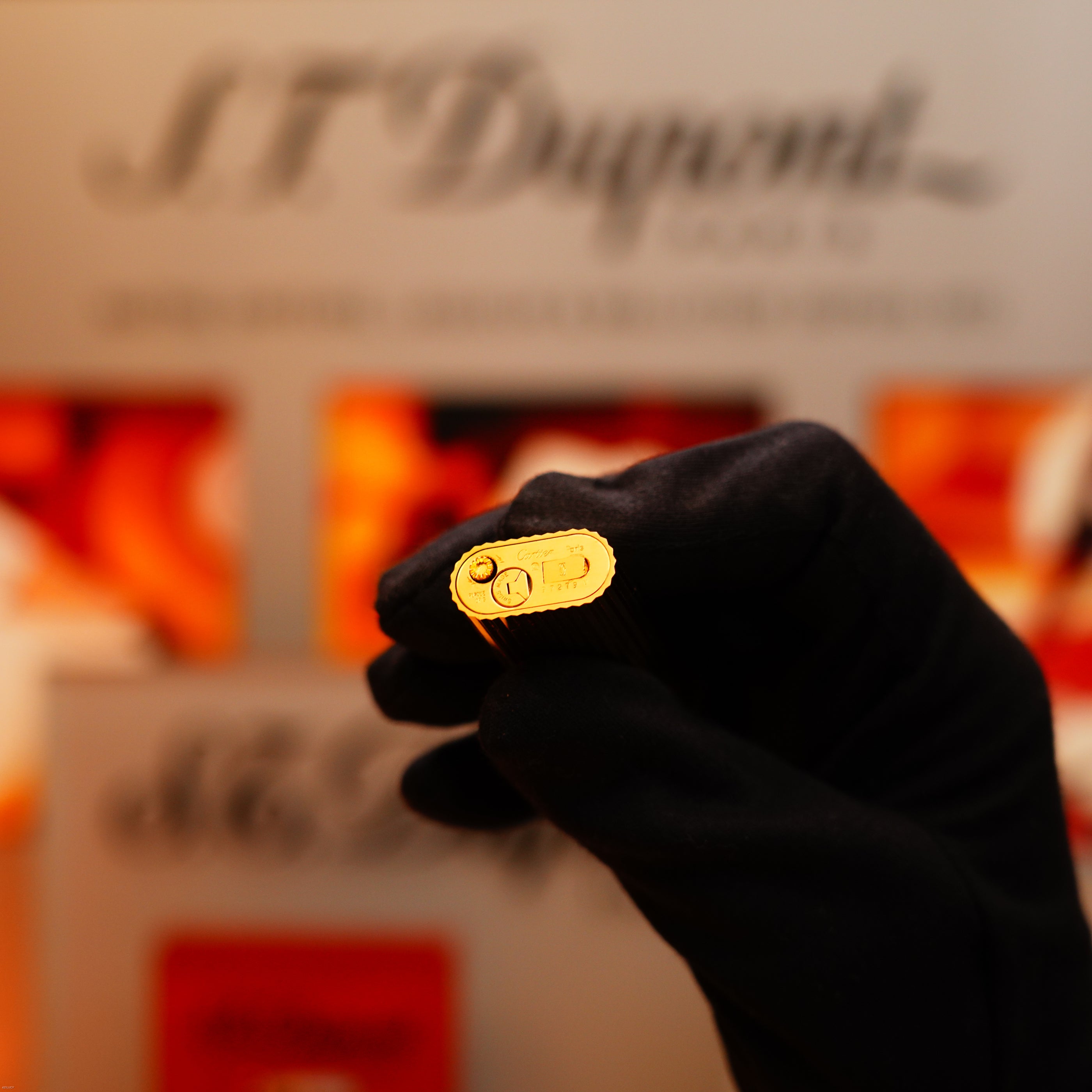 A gloved hand holds a Vintage 1980 Cartier Le Must lighter with a heavy 24k gold finish and vertical lines, the brand name engraved on the bottom, with a blurred background displaying the Cartier logo.