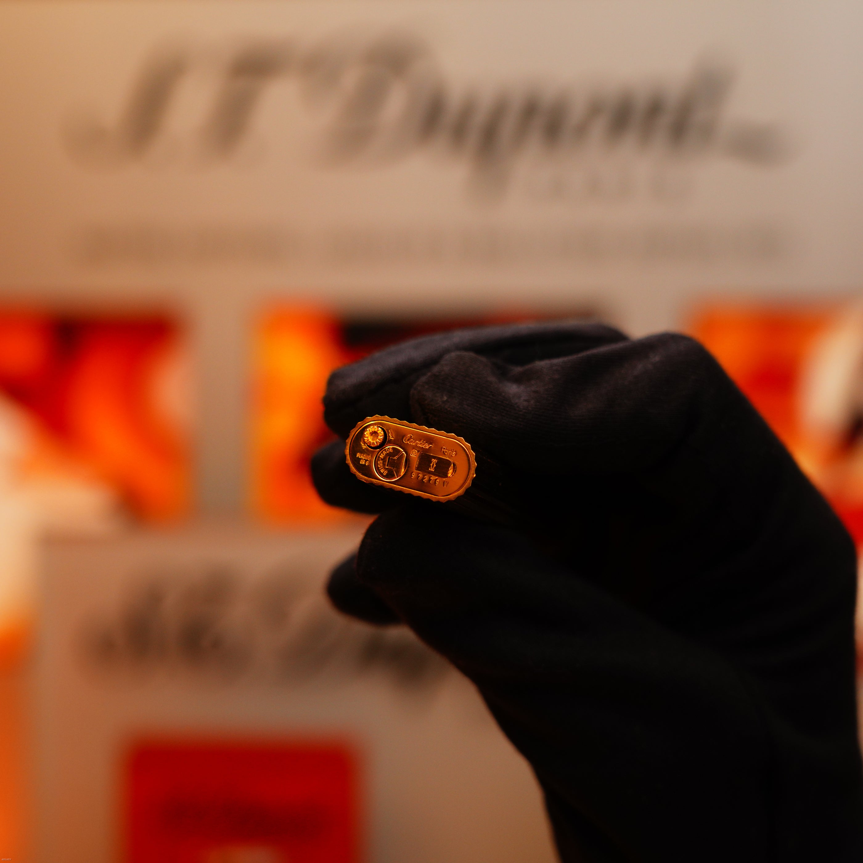 A gloved hand holds the Vintage 1980 Cartier Le Must Oval Lighter with heavy 24k gold finish and engraved vertical lines, in front of a blurred Cartier display background.