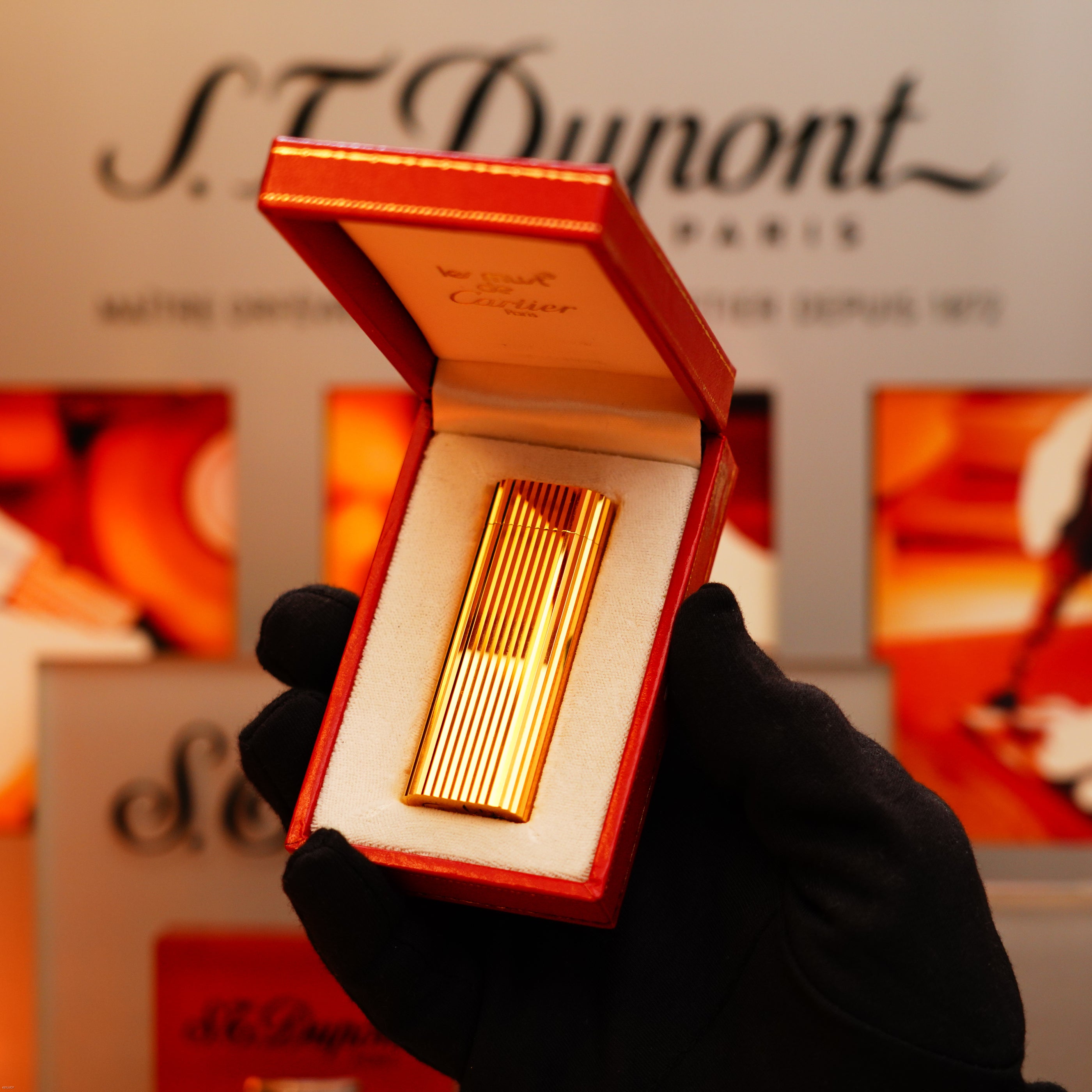 A hand in a black glove holds an open red box displaying a Vintage 1980 Cartier Vertical line Heavy 24k Gold Finish Oval Le Must Lighter, with Cartier-branded packaging in the background.