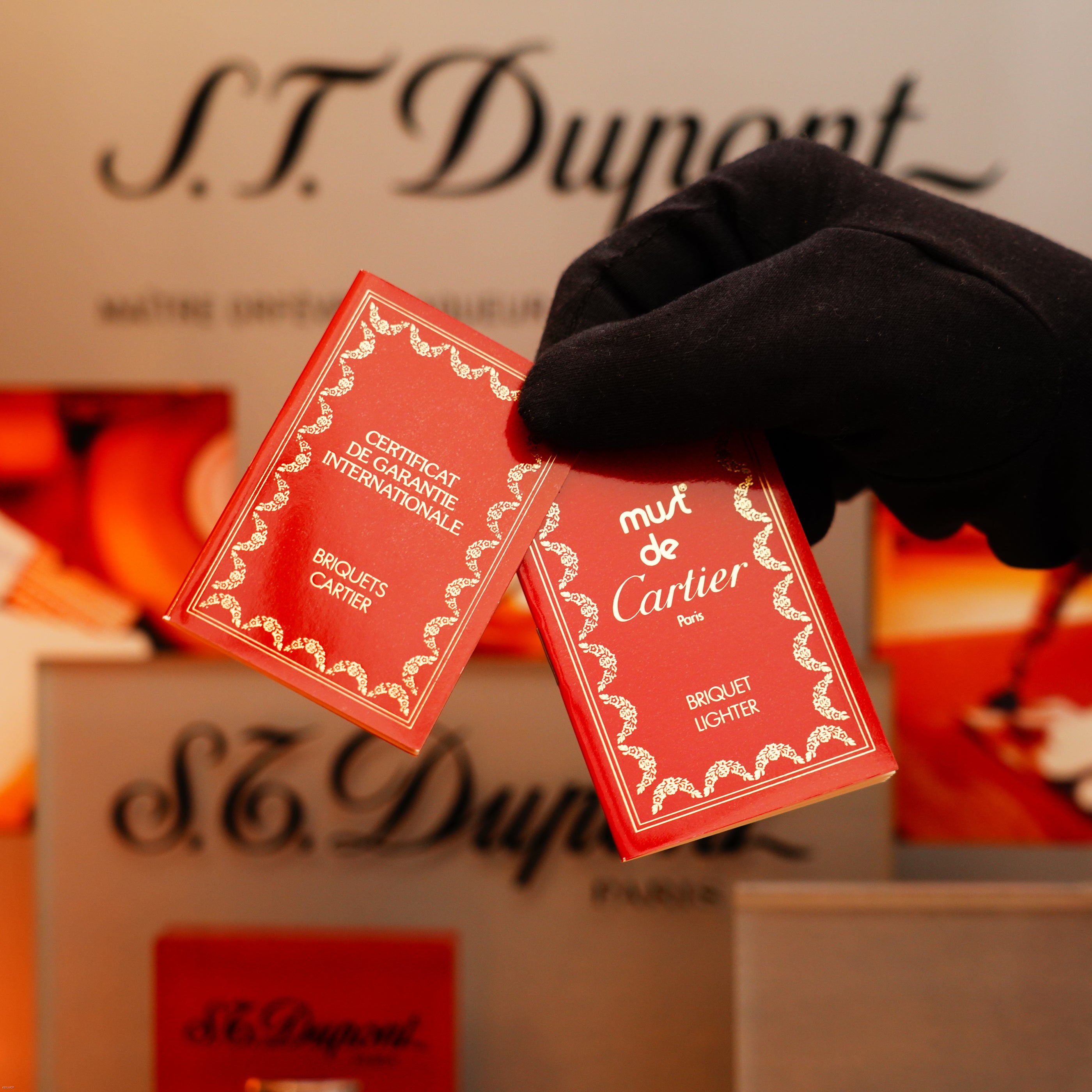 A gloved hand holds two red certificates—one marked "Certificat de Garantie Internationale S.T. Dupont" and the other "Must de Cartier Paris"—in front of packaging for a Vintage 1980 Cartier Le Must Oval Lighter with heavy 24k gold finish.