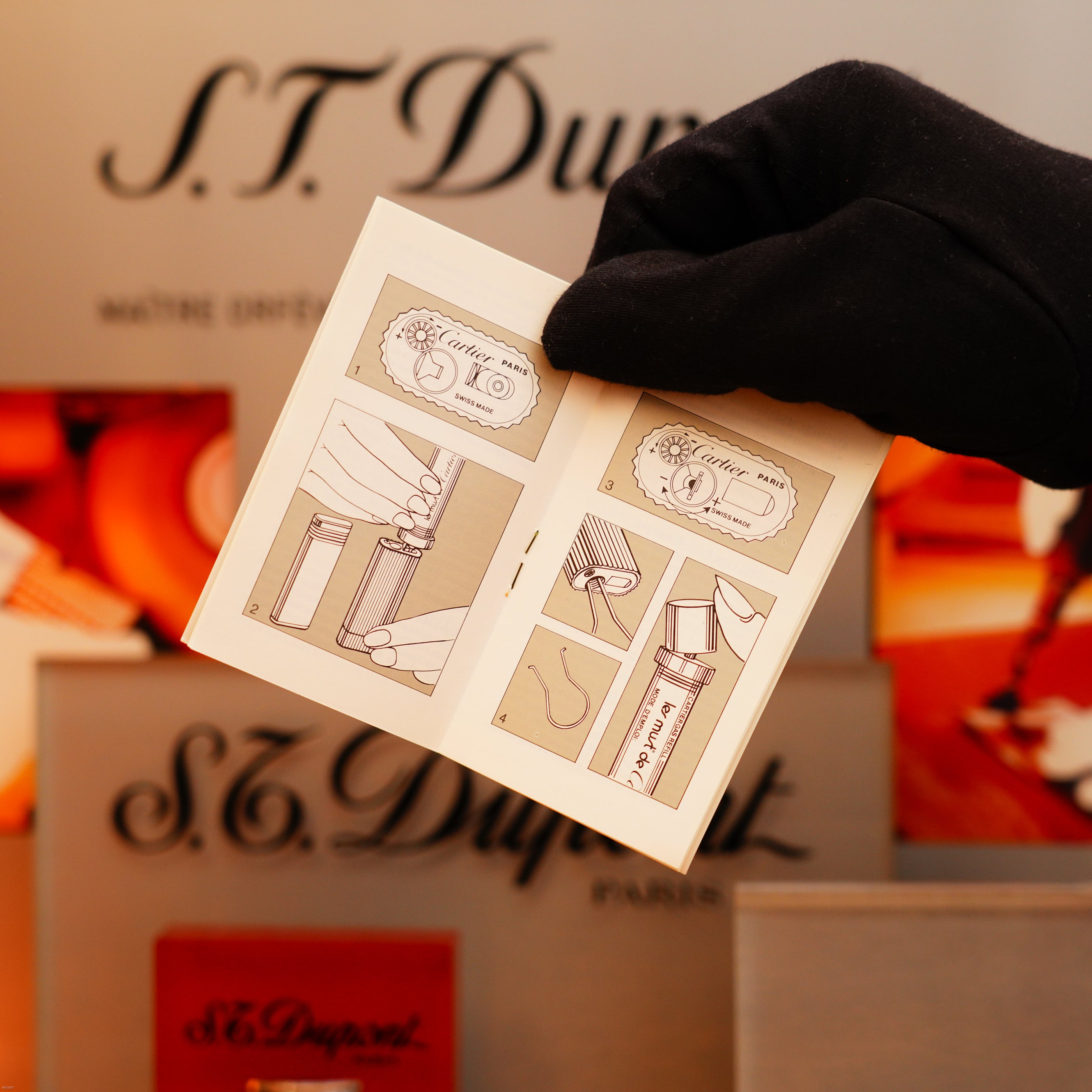 A gloved hand holds a small instruction booklet with illustrations for using a Vintage 1980 Cartier Vertical line Heavy 24k Gold Finish Oval Le Must Lighter, against a background of Cartier branded boxes.