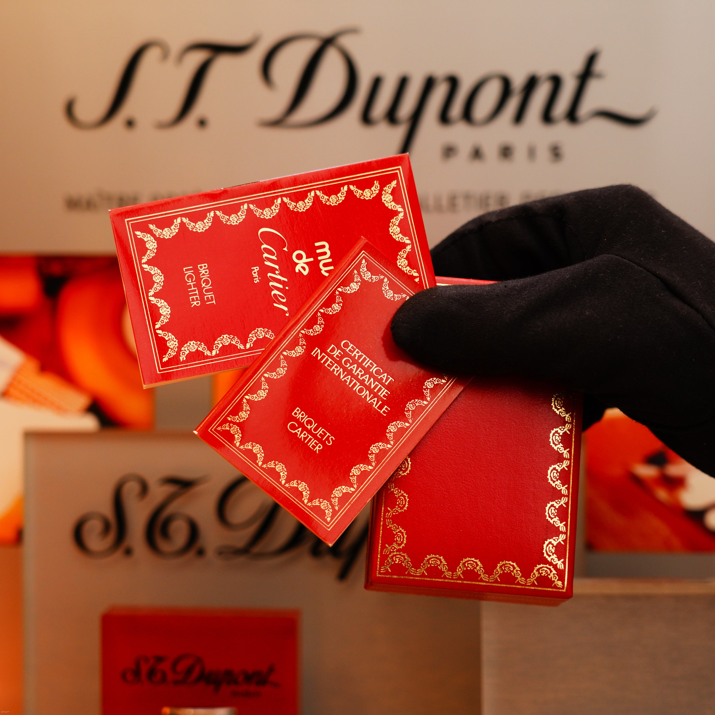 A gloved hand holds three red Cartier booklets with gold detailing in front of a blurred display, referencing the Vintage 1980 Cartier Vertical Line Heavy 24k Gold Finish Oval Le Must Lighter.