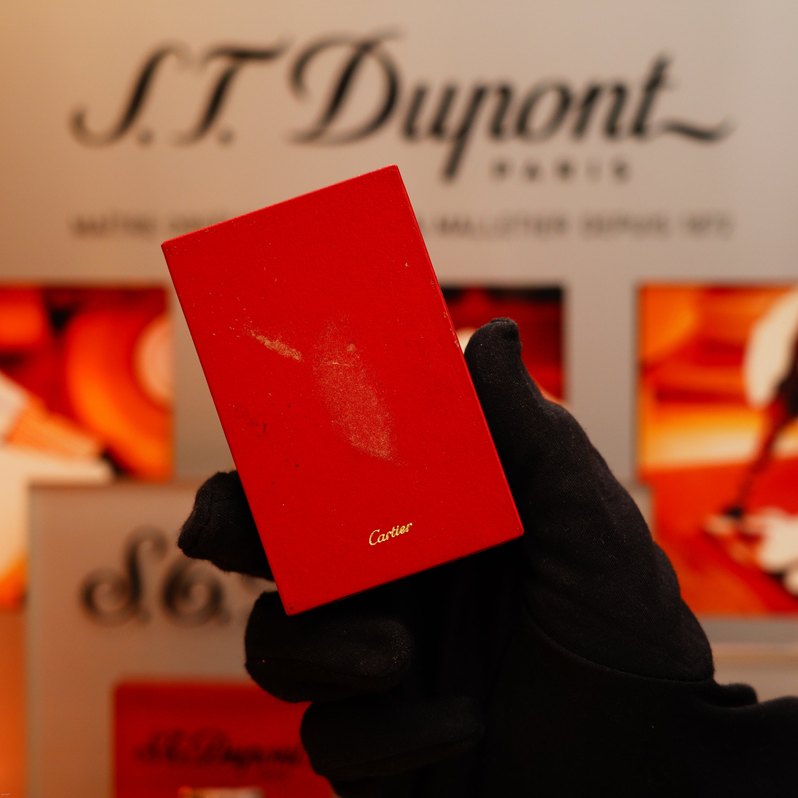A gloved hand holds a Vintage 1980 Cartier Vertical Line Heavy 24k Gold Finish Oval Le Must Lighter with a scratch, in front of an S.T. Dupont Paris display.