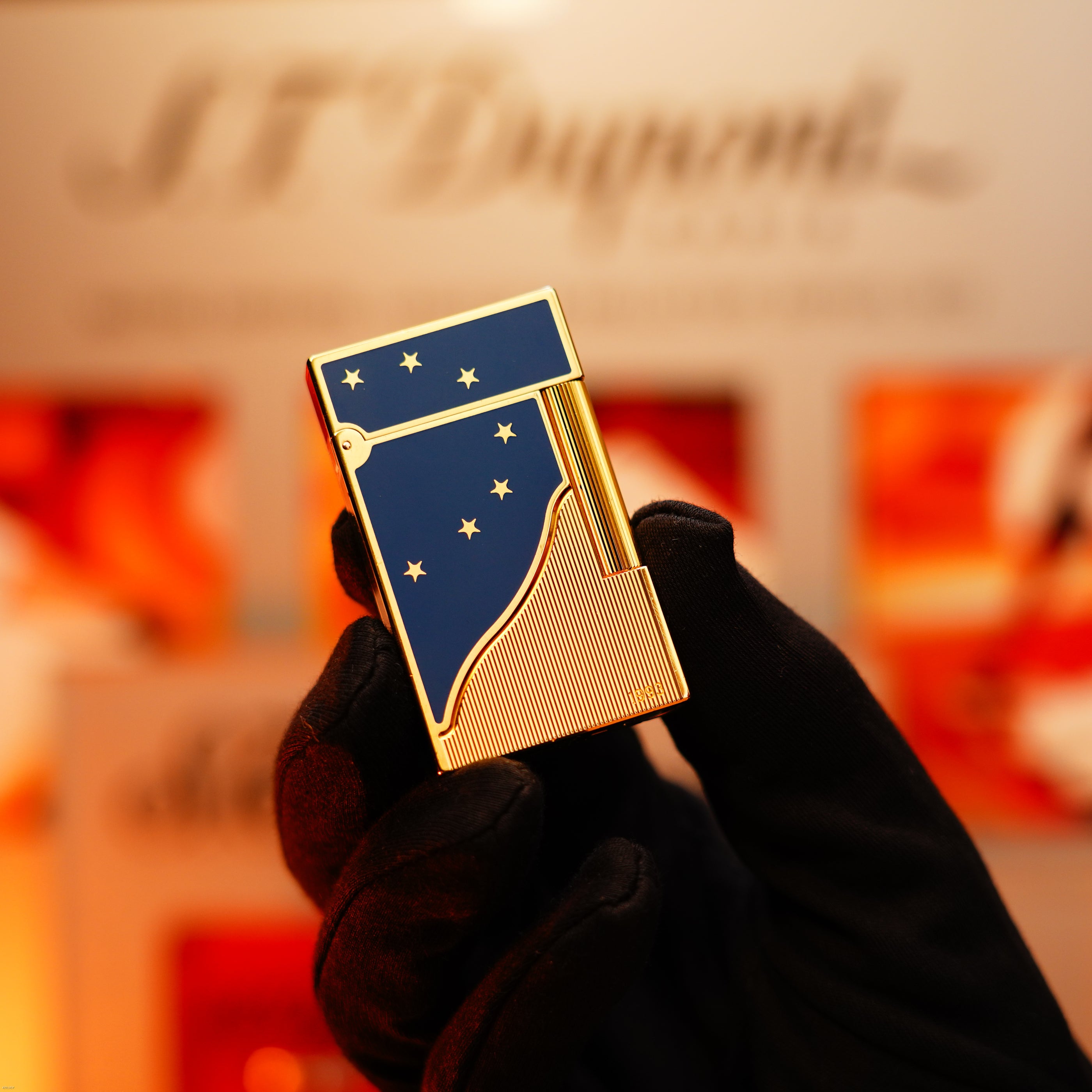 A gloved hand holds a Lucy Lighter Vintage 1993 St Dupont Europe Rare 24k Gold Finish Blue Lacquer Limited Edition Ligne 2 lighter with a star pattern, set against a blurred background.