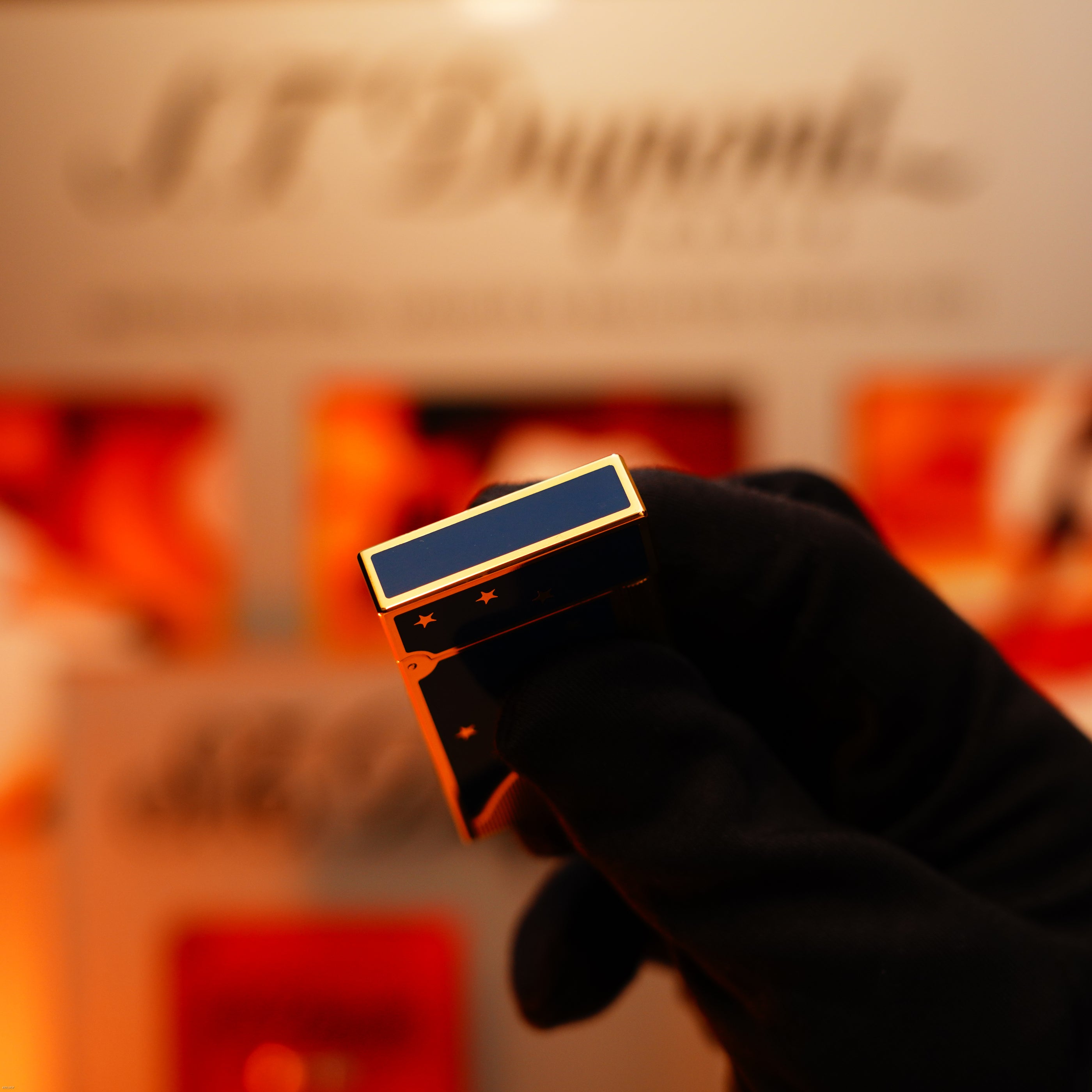 A gloved hand holds a Lucy Lighter Vintage 1993 St Dupont Europe Rare 24k Gold Finish Blue Lacquer Limited Edition Ligne 2 Lighter in front of a blurred S.T. Dupont display background.