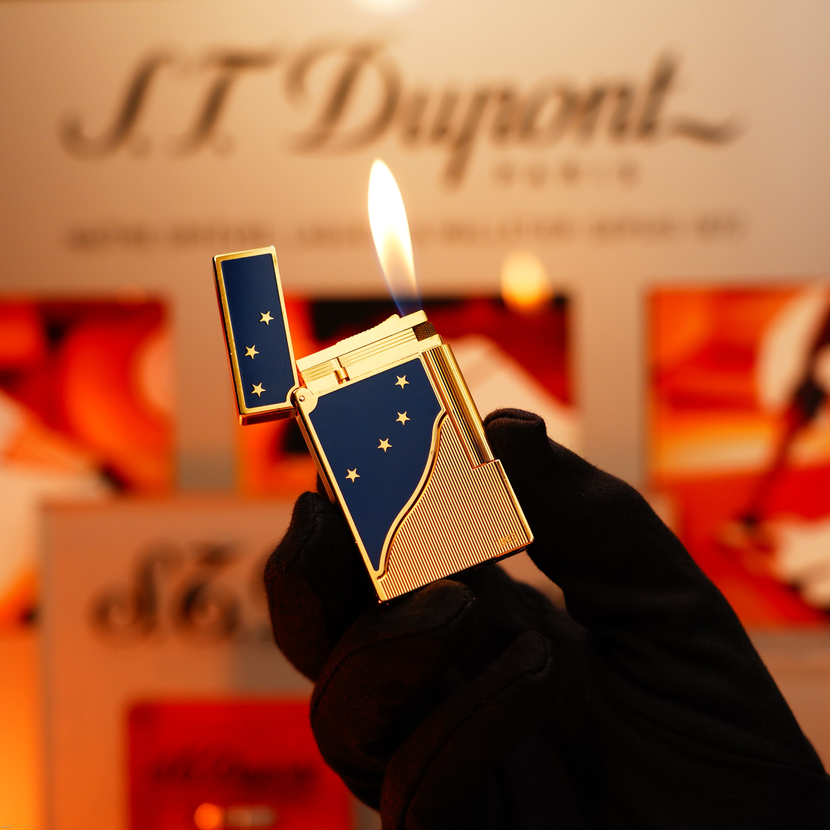 A gloved hand holds a Lucy Lighter Vintage 1993 St Dupont Europe Rare 24k Gold Finish Blue Lacquer Limited Edition Ligne 2 Lighter, its flame alight against a blurred branded background.