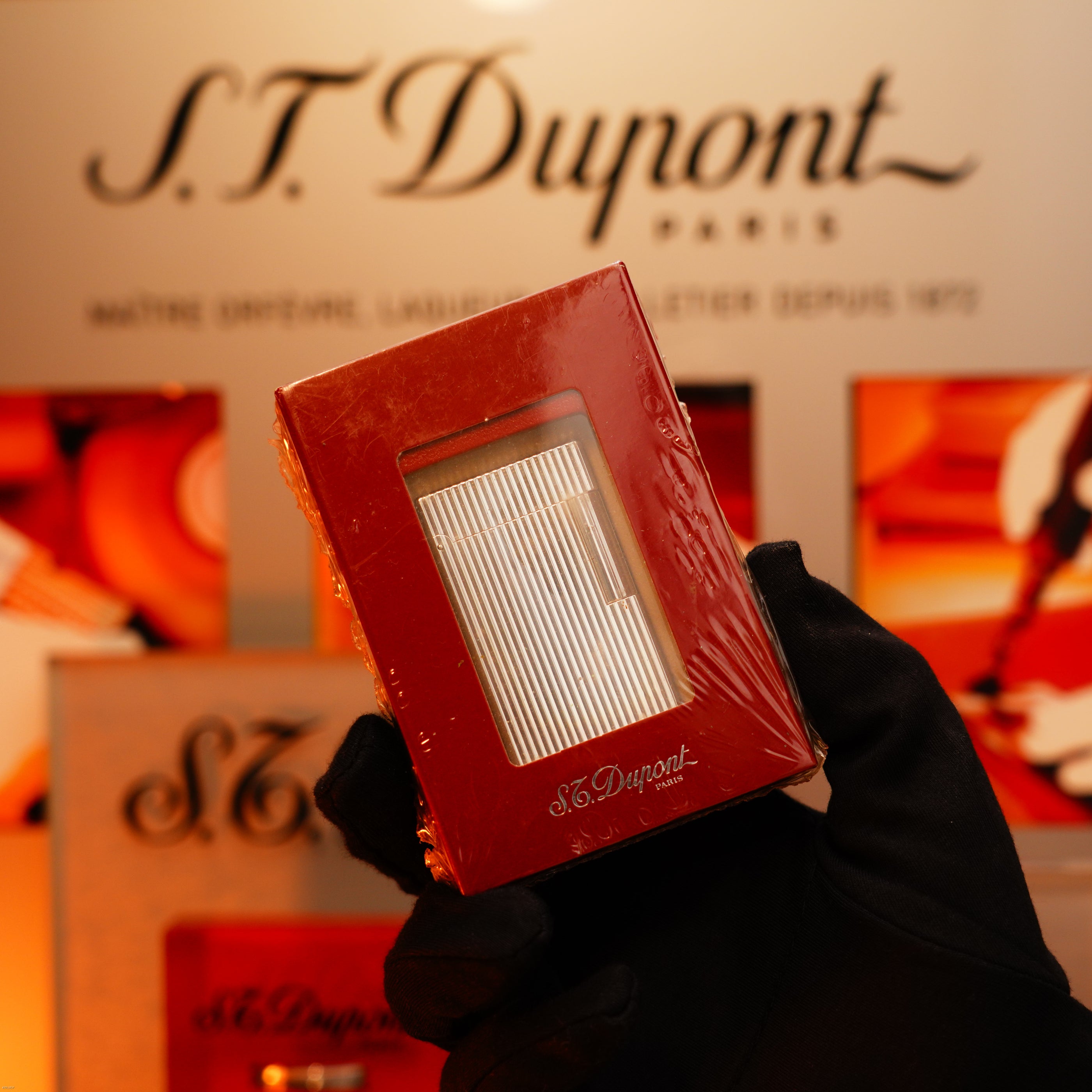 A gloved hand holds a boxed Vintage 1990 St Dupont Heavy Silver iconic Vertical ligne rare Sealed Lighter by Lucy Lighter in front of a blurred branded display, highlighting its timeless style and quality assurance.