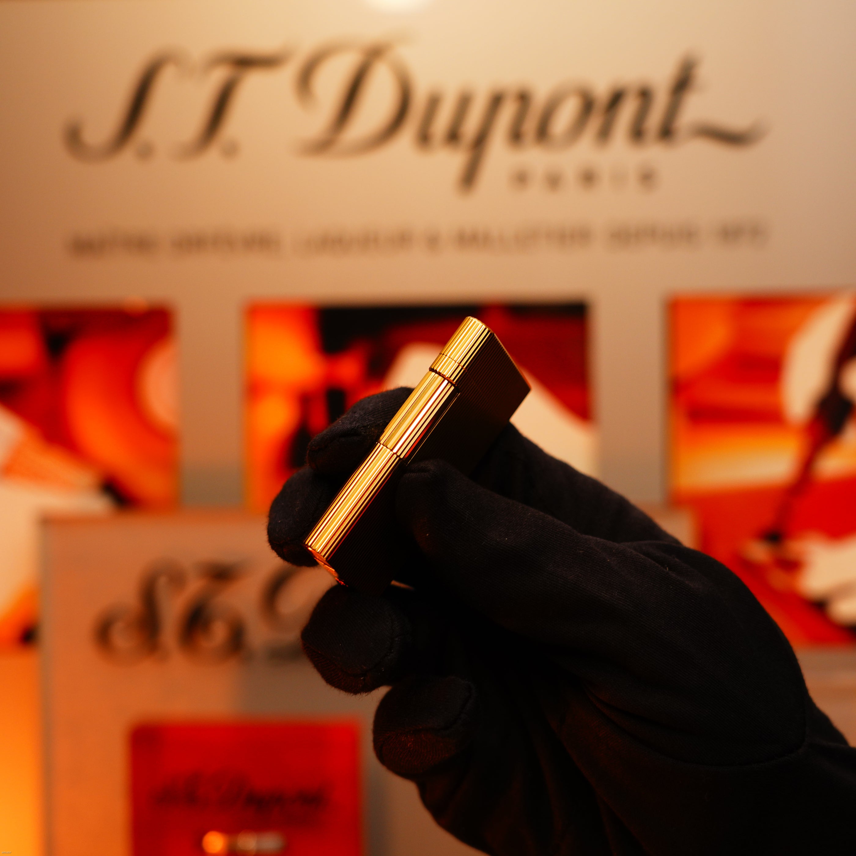 A gloved hand holds a Lucy Lighter Vintage 1970 Heavy 24k Gold Finish Caran d'Ache Limited Madison Swiss Lighter—extremely rare—with a blurred brand display in the background. Includes a 12-month warranty against functional issues.