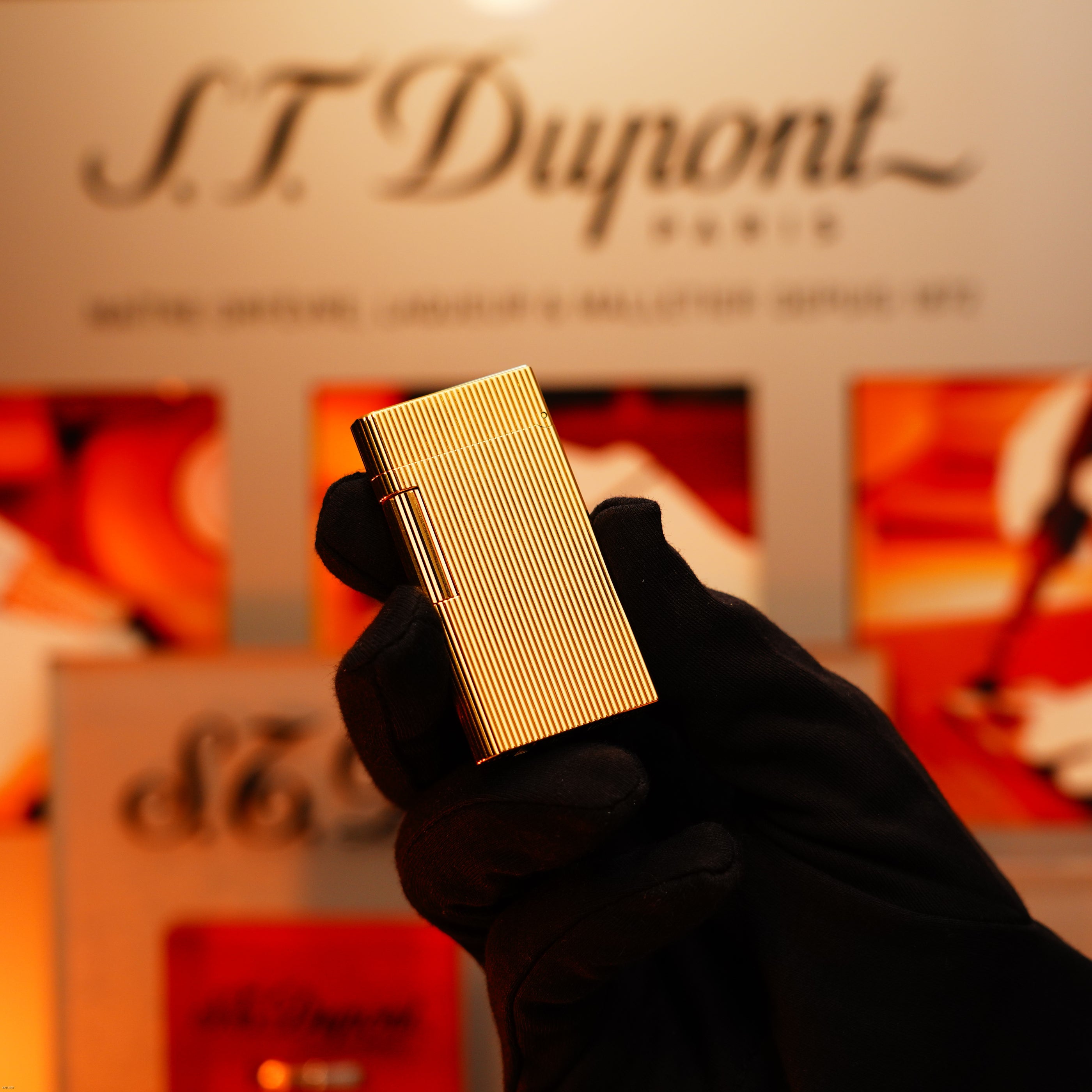 A gloved hand holds a Vintage 1970 Heavy 24k Gold Finish Caran d'Ache Limited Madison Lighter by Lucy Lighter in front of a blurred background with logo and packaging; comes with a 12-month warranty for functional issues.
