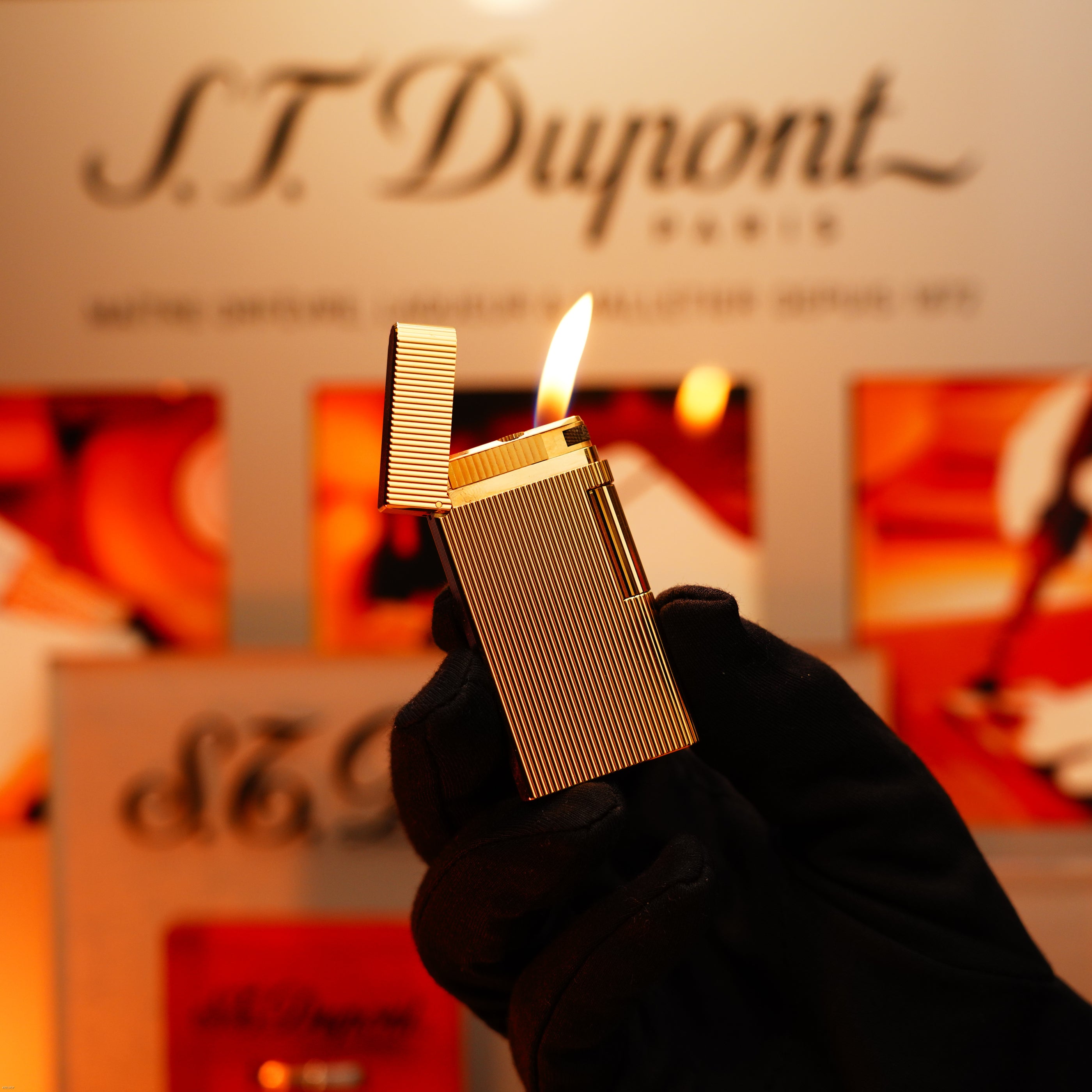 A gloved hand holds the Vintage 1970 Heavy 24k Gold Finish Caran d'Ache Madison Swiss Lighter by Lucy Lighter, its flame lit before a blurred display—an extremely rare luxury piece with a 12-month warranty for functional issues.