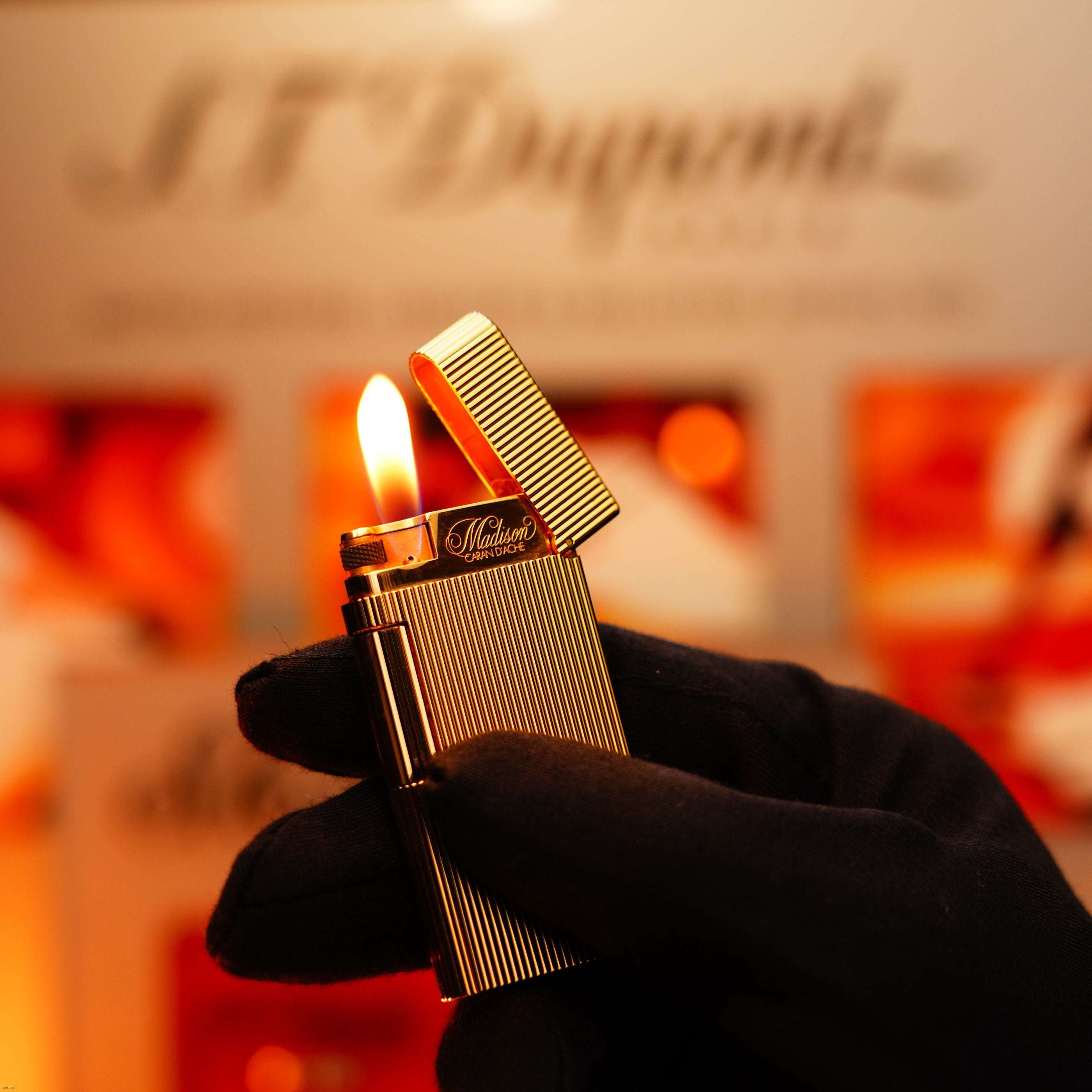 A gloved hand holds a lit Vintage 1970 Heavy 24k Gold Finish Caran d'ache Limited Madison Lighter by Lucy Lighter, its lid open, against a blurred background with the Lucy Lighter logo. Includes a 12-month warranty for functional issues.