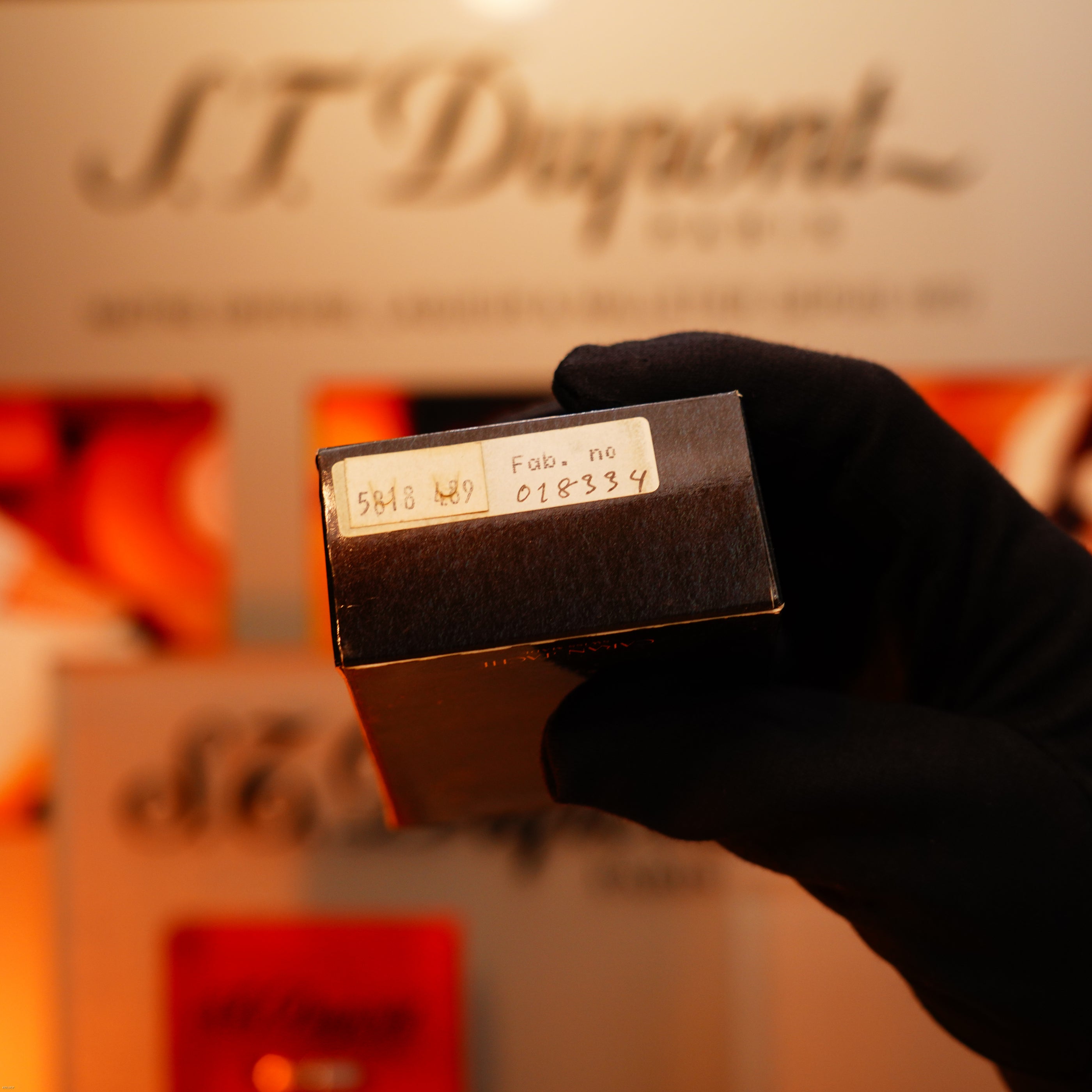 A gloved hand holds a black box labeled "5848 AG" and "Fab. no 018334" before a blurred display, showcasing the rare Lucy Lighter Vintage 1970 Heavy 24k Gold Finish Caran d'Ache Madison Swiss Lighter with a 12-month functional warranty.