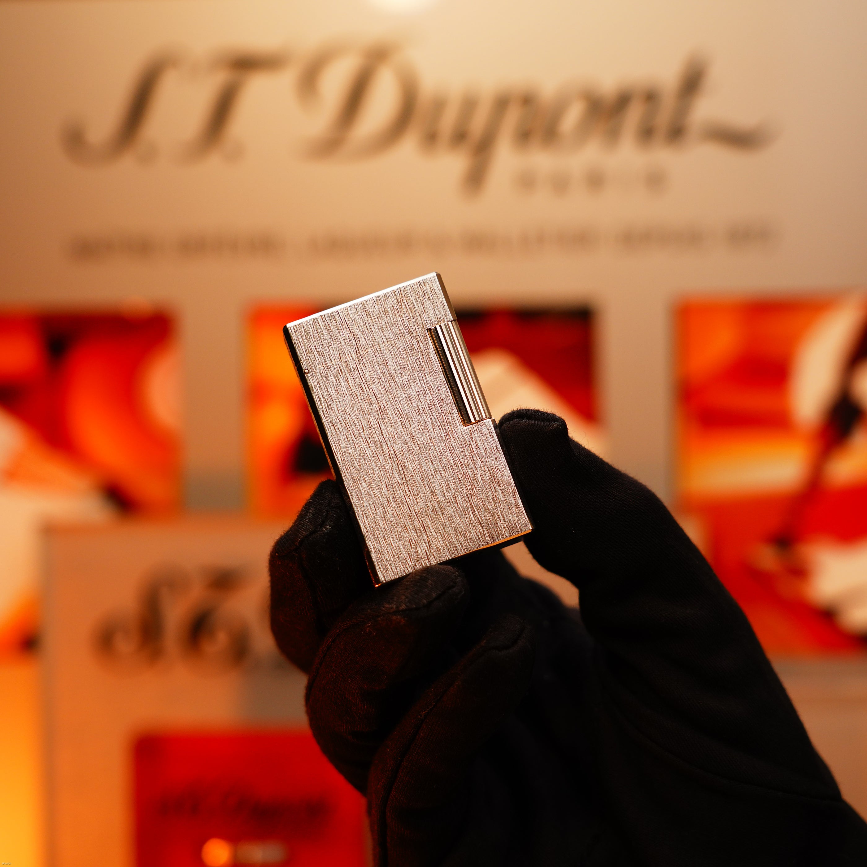 A gloved hand holds a Vintage 1980 St Dupont Heavy Silver Finish Ligne 1 Large Rare Pattern Lighter by Lucy Lighter, with a blurred sign and warm-toned background visible behind it.