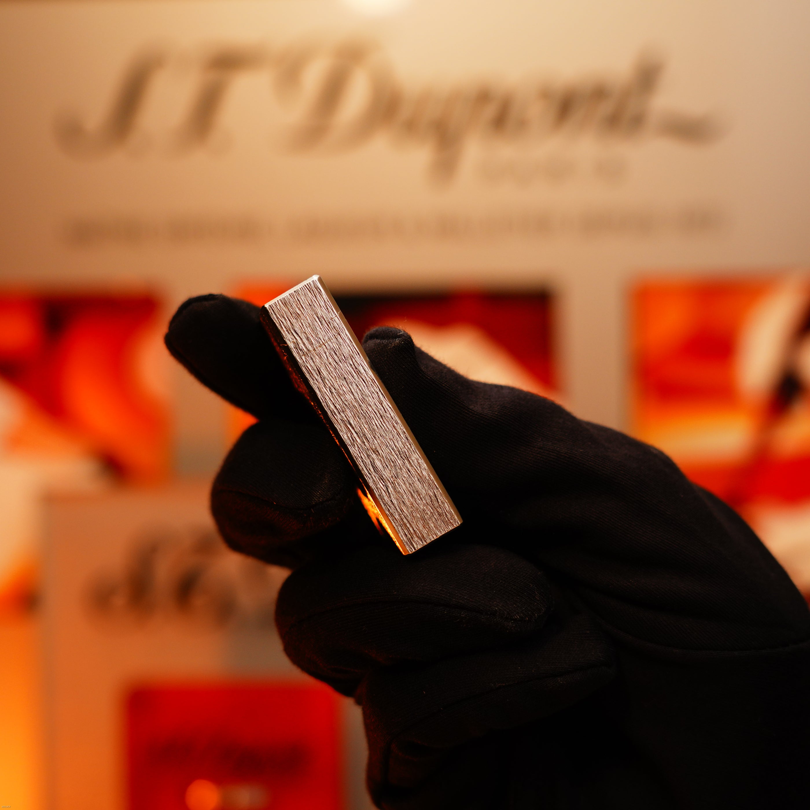 A gloved hand holds a Vintage 1980 St Dupont Heavy Silver Finish Ligne 1 Large Rare Pattern Lighter by Lucy Lighter upright before a blurred S.T. Dupont display.