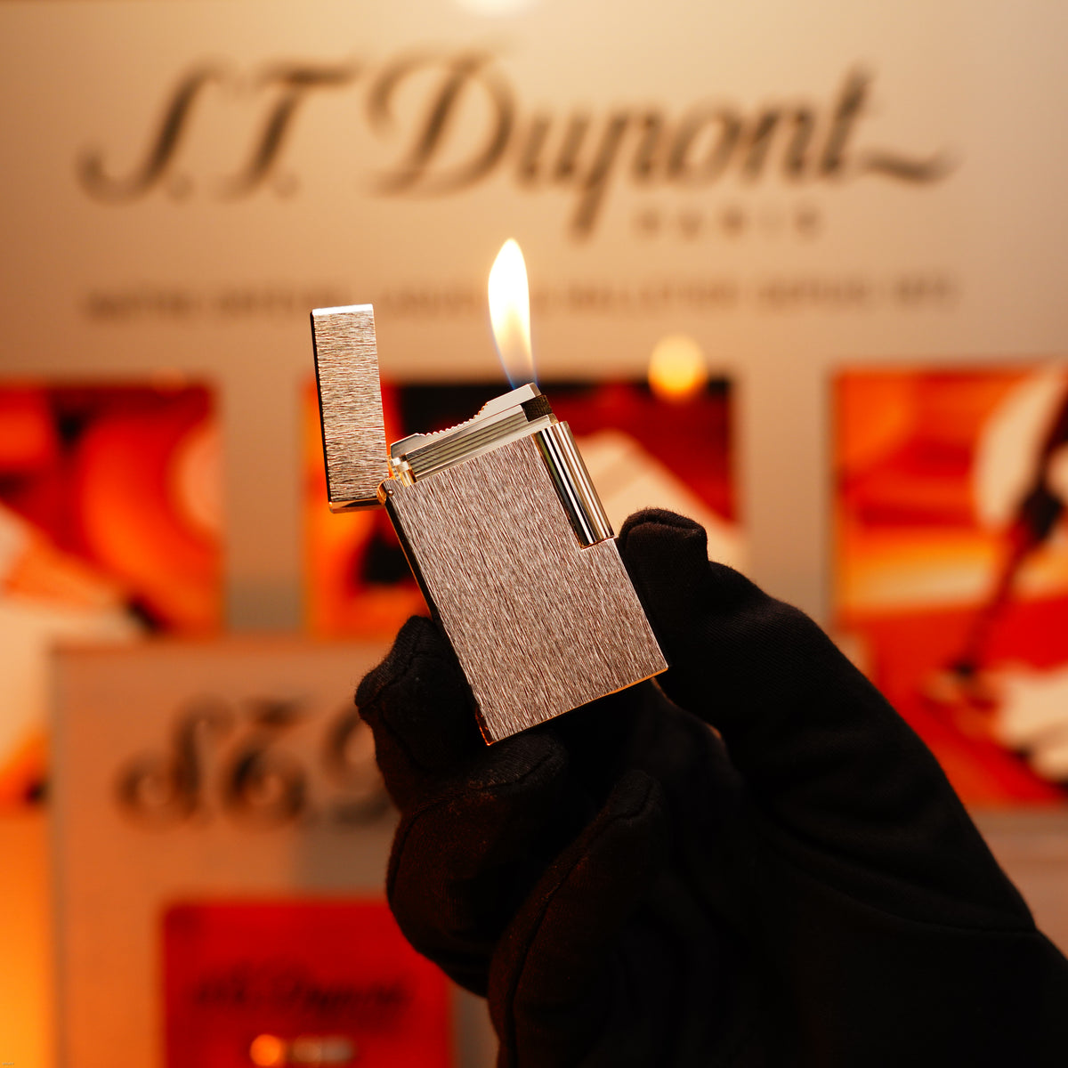 A gloved hand holds a lit Lucy Lighter Vintage 1980 St Dupont Heavy Silver Finish Ligne 1 Large Rare Pattern Lighter; its flame stands out against the blurred background with logo and packaging, emphasizing its luxury and quality.