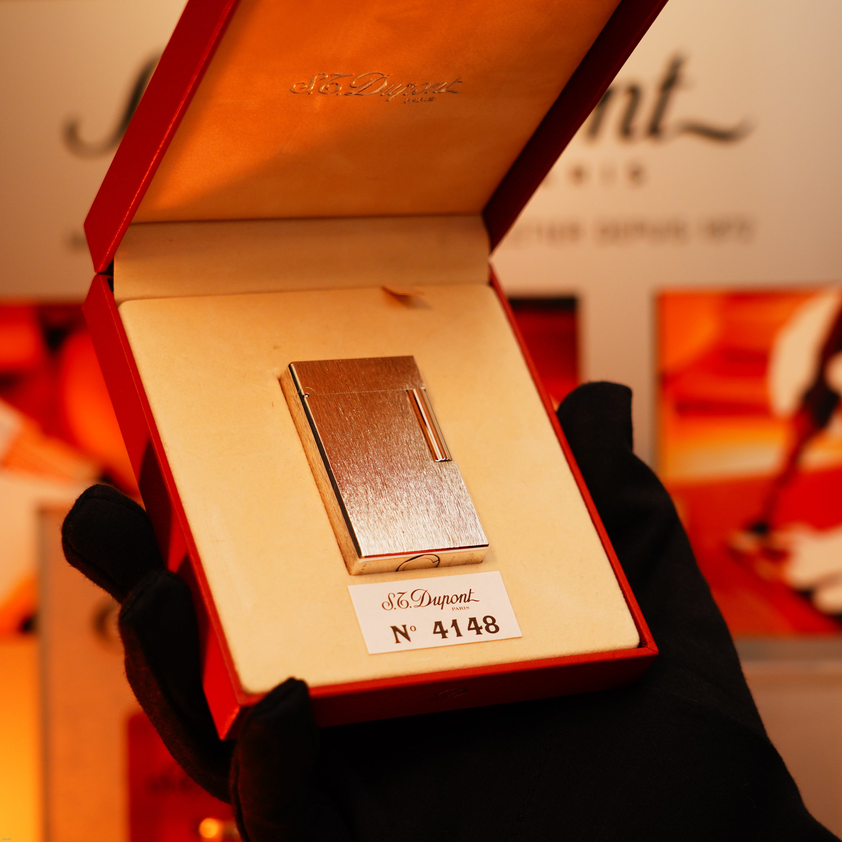 A gloved hand holds an open red box displaying a Lucy Lighter Vintage 1980 St Dupont Heavy Silver Finish Ligne 1 Large size Rare Pattern Lighter, set against a blurred background.