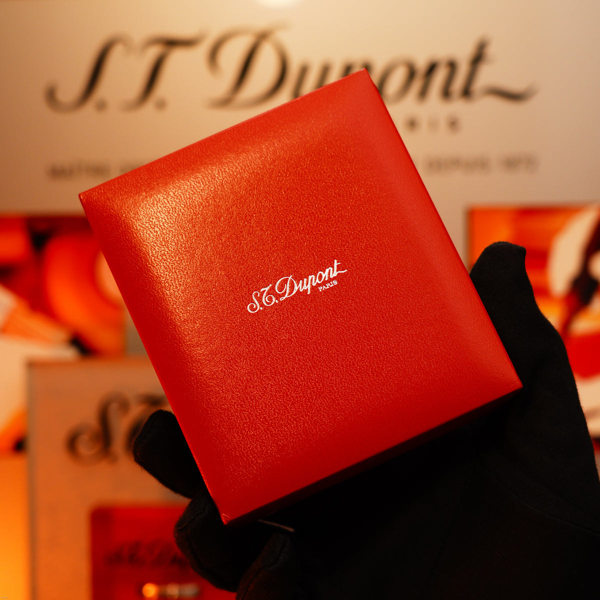 A gloved hand holds a Vintage 1980 Lucy Lighter Heavy Silver Finish Ligne 1 Large Rare Pattern Lighter box, the Lucy Lighter logo clear in white—showcasing luxury and quality. The logo also appears in the blurred background.