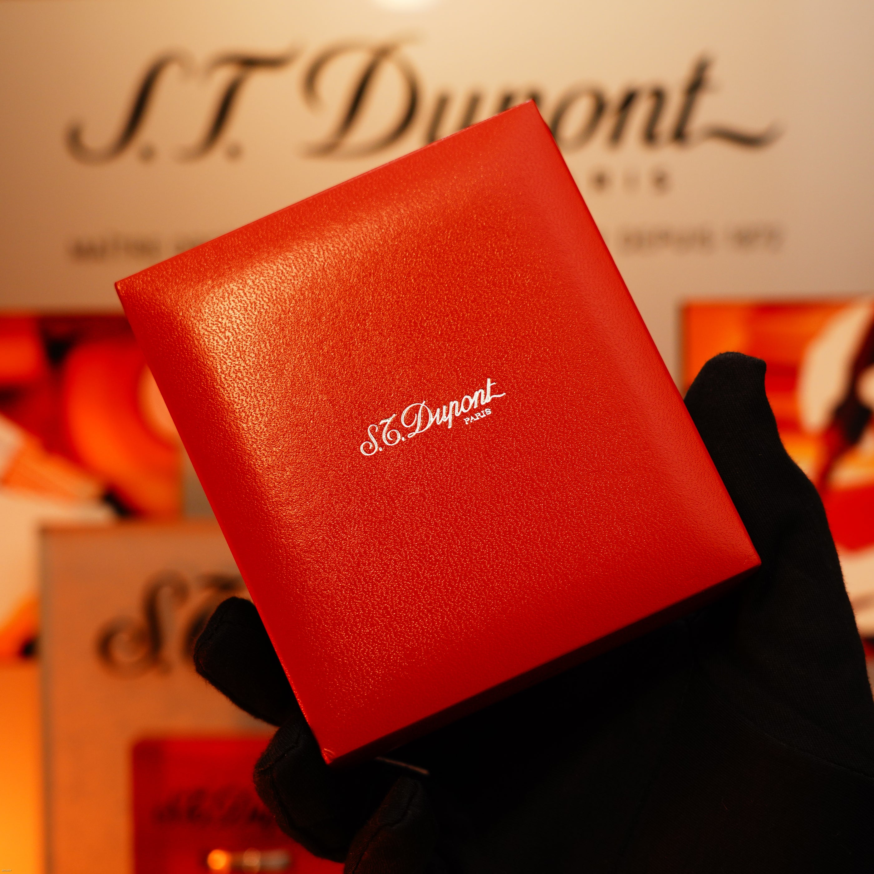 A gloved hand holds a Vintage 1980 Lucy Lighter Heavy Silver Finish Ligne 1 Large Rare Pattern Lighter box, the Lucy Lighter logo clear in white—showcasing luxury and quality. The logo also appears in the blurred background.