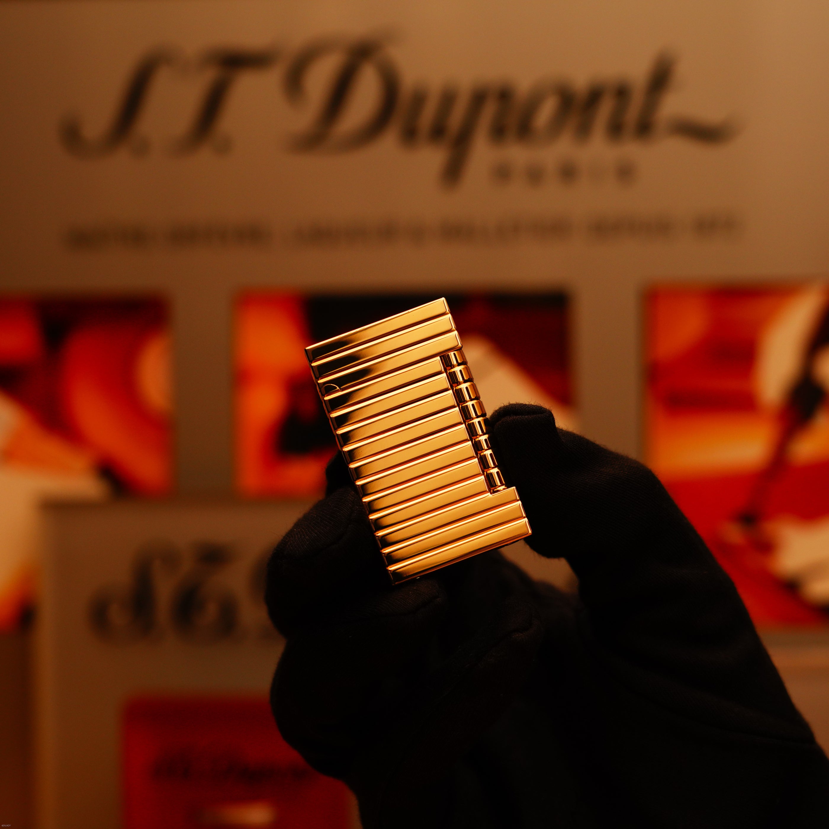 A gloved hand holds the ultra rare Vintage 1970 ST Dupont Lighter—Solid 18K Gold Masterpiece by Lucy Lighter, set against a blurred background with the S.T. Dupont Paris logo, assuring quality and exclusivity.