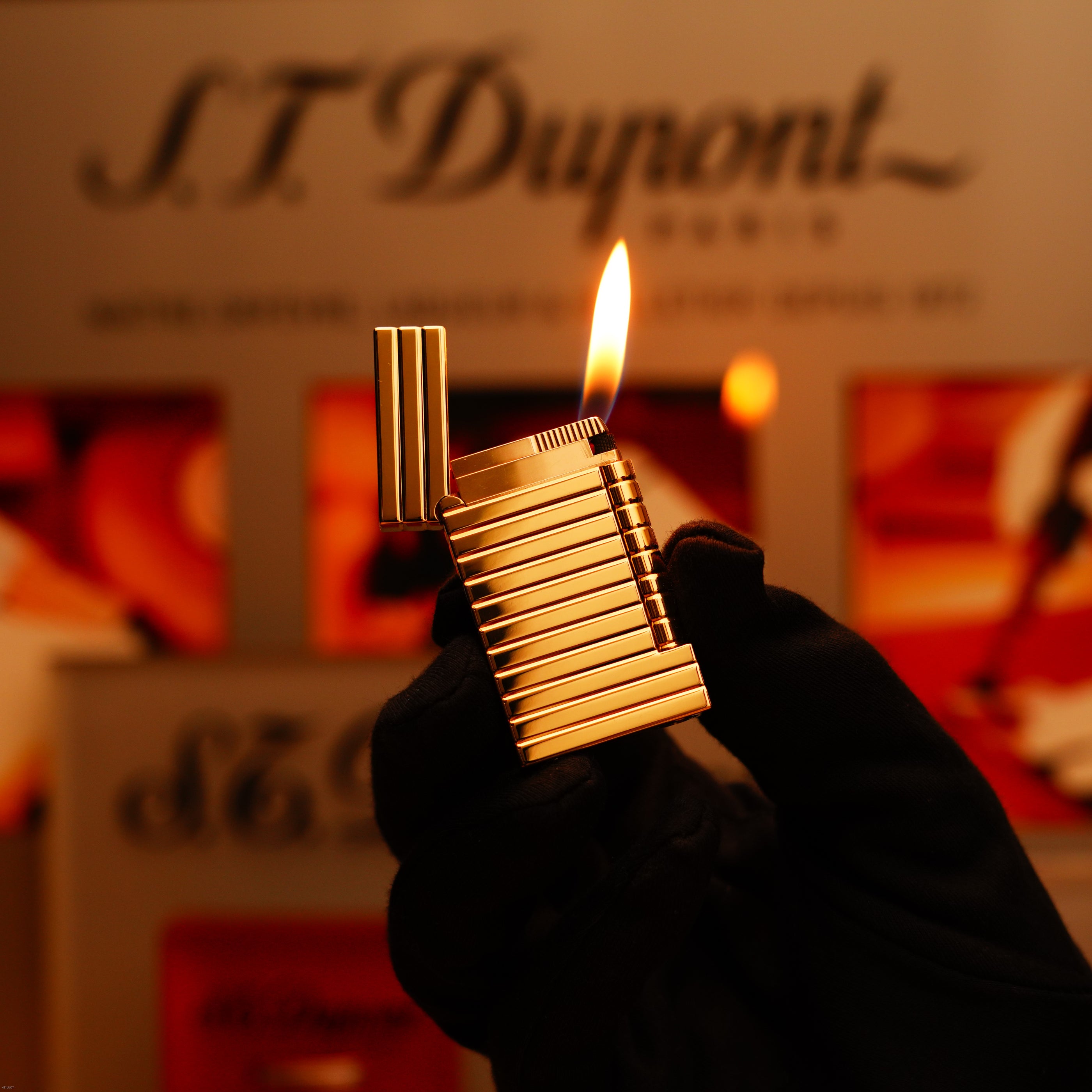 A gloved hand holds a lit Lucy Lighter Vintage 1970 Ultra Rare 1/1 ST Dupont Lighter – Solid 18K Gold Masterpiece, with its flame against a blurred branded background.