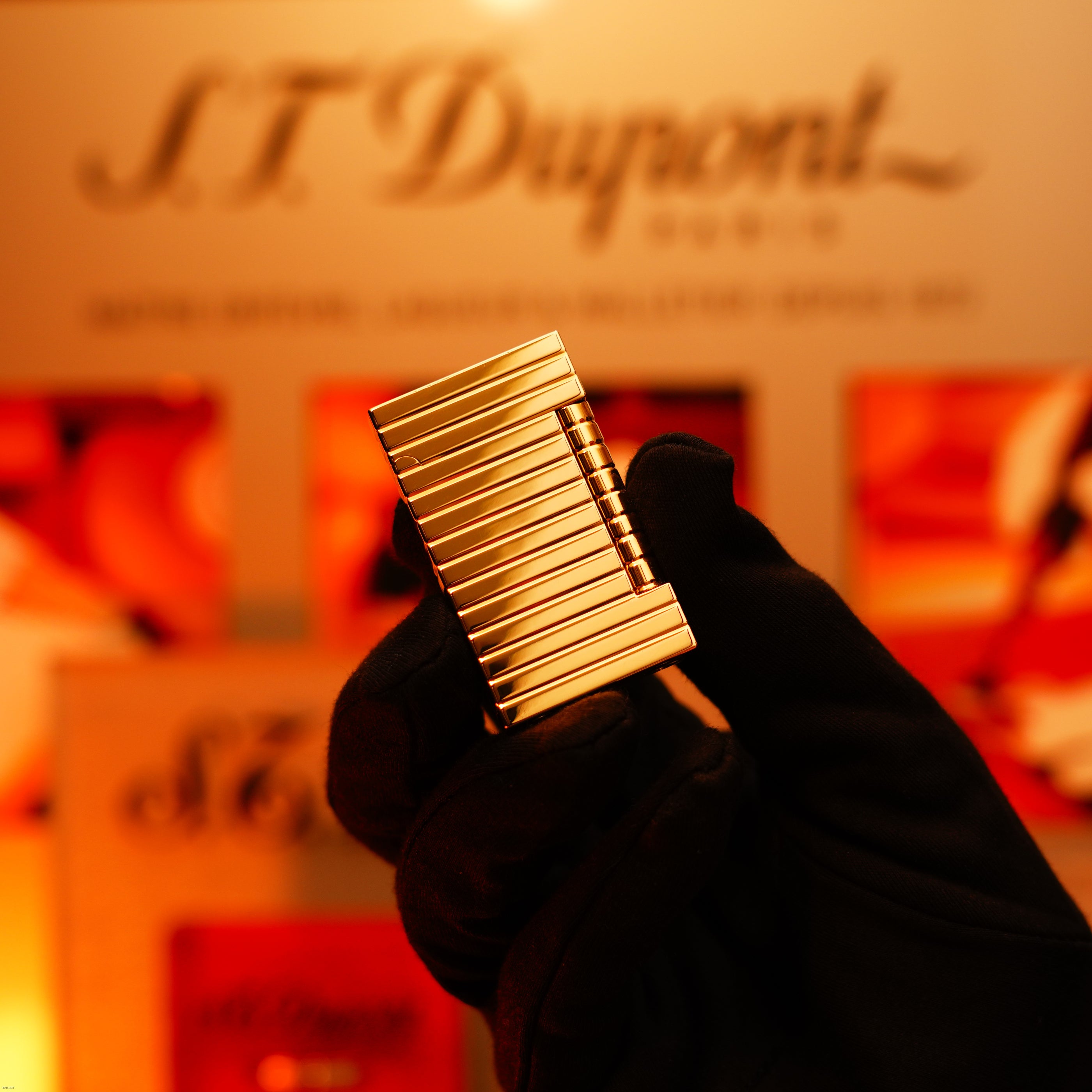 A gloved hand displays the ultra-rare Vintage 1970 ST Dupont Lighter by Lucy Lighter, crafted in solid 18K gold, with a blurred background showing the logo and packaging, emphasizing its exclusivity and refined craftsmanship.