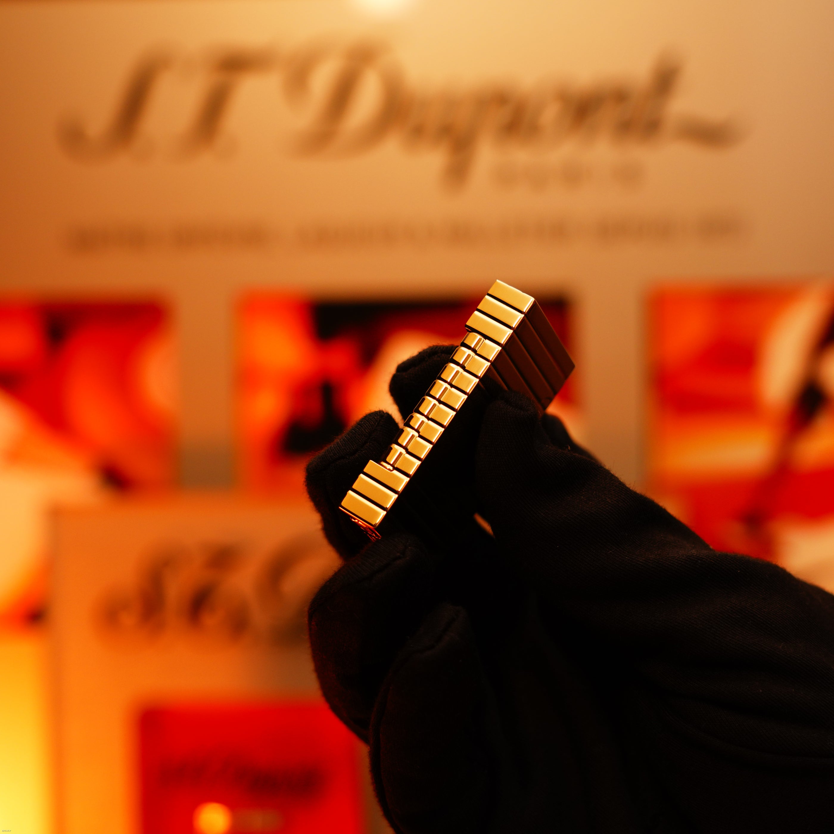 A gloved hand holds the Vintage 1970 Ultra Rare 1/1 ST Dupont Lighter by Lucy Lighter, a solid 18K gold masterpiece with ridged edges; blurred branding in the background emphasizes its quality and luxury.