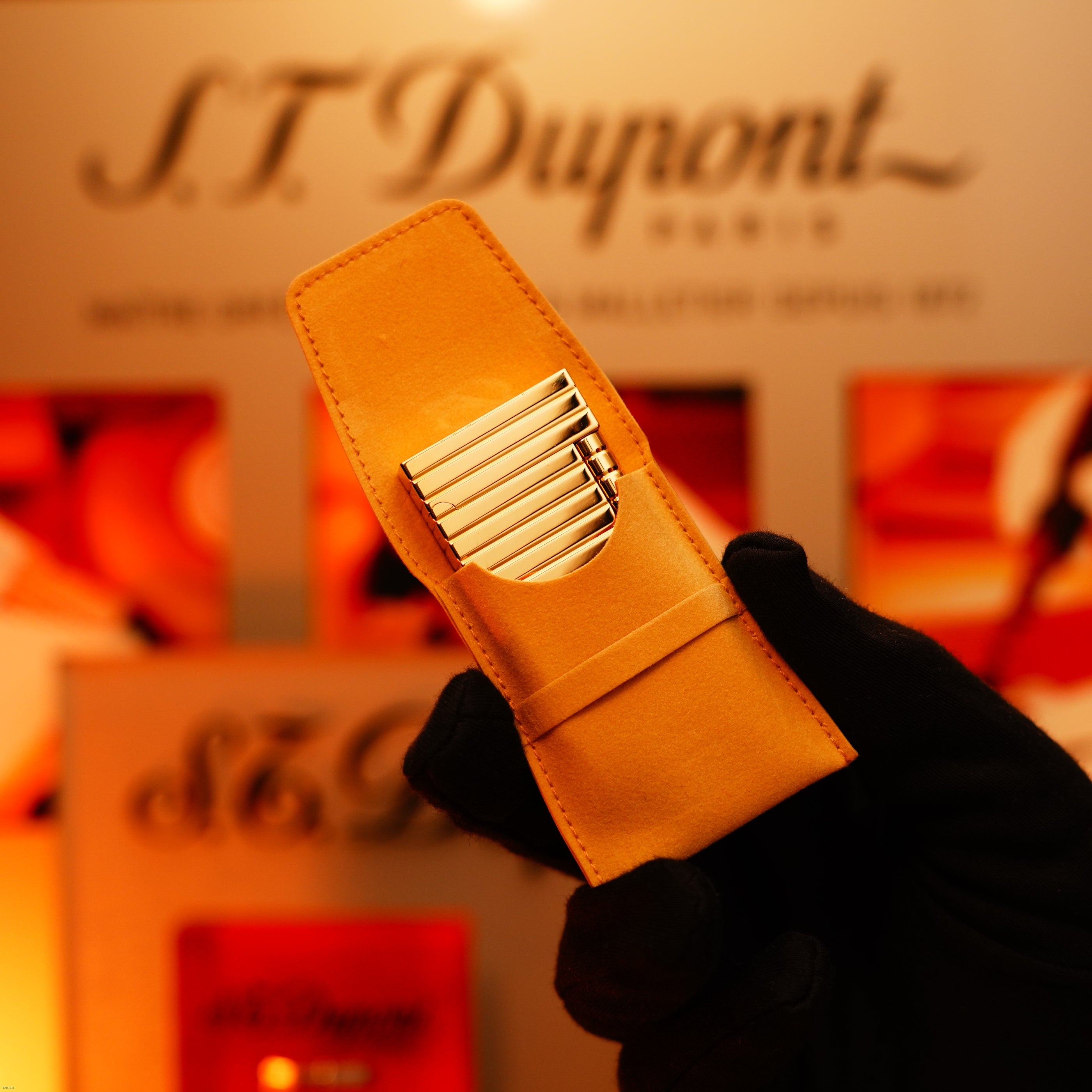 A gloved hand holds a tan leather case with a Vintage 1970 Ultra Rare 1/1 ST Dupont Lighter – Solid 18K Gold Masterpiece by Lucy Lighter, with an S.T. Dupont Paris sign visible in the blurred background.