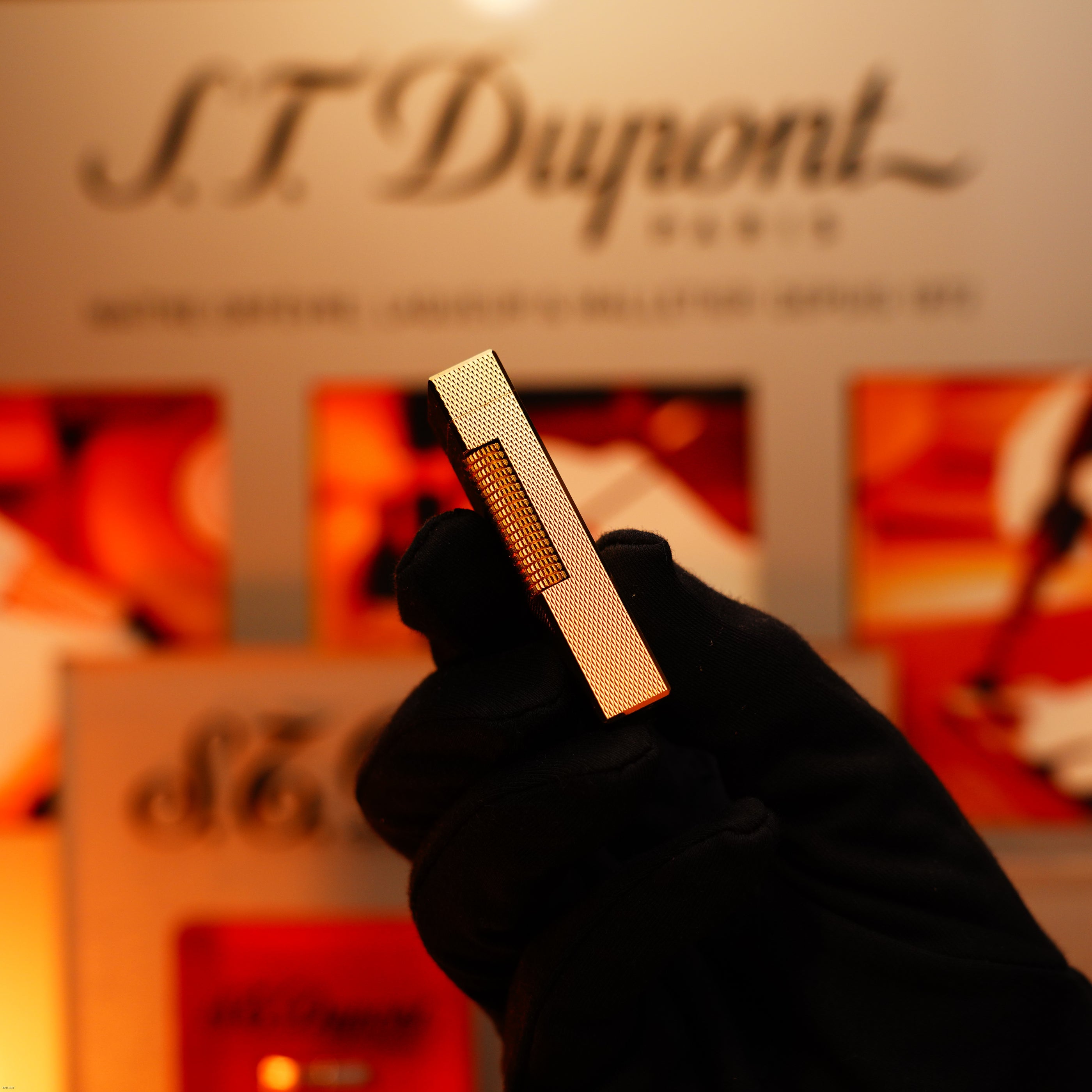 A hand in a black glove holds a vintage 1975 Dunhill 24k Heavy Gold Finish Barley Pattern early version lighter by Lucy Lighter, against a blurred background with the Lucy Lighter logo and warm-toned displays.