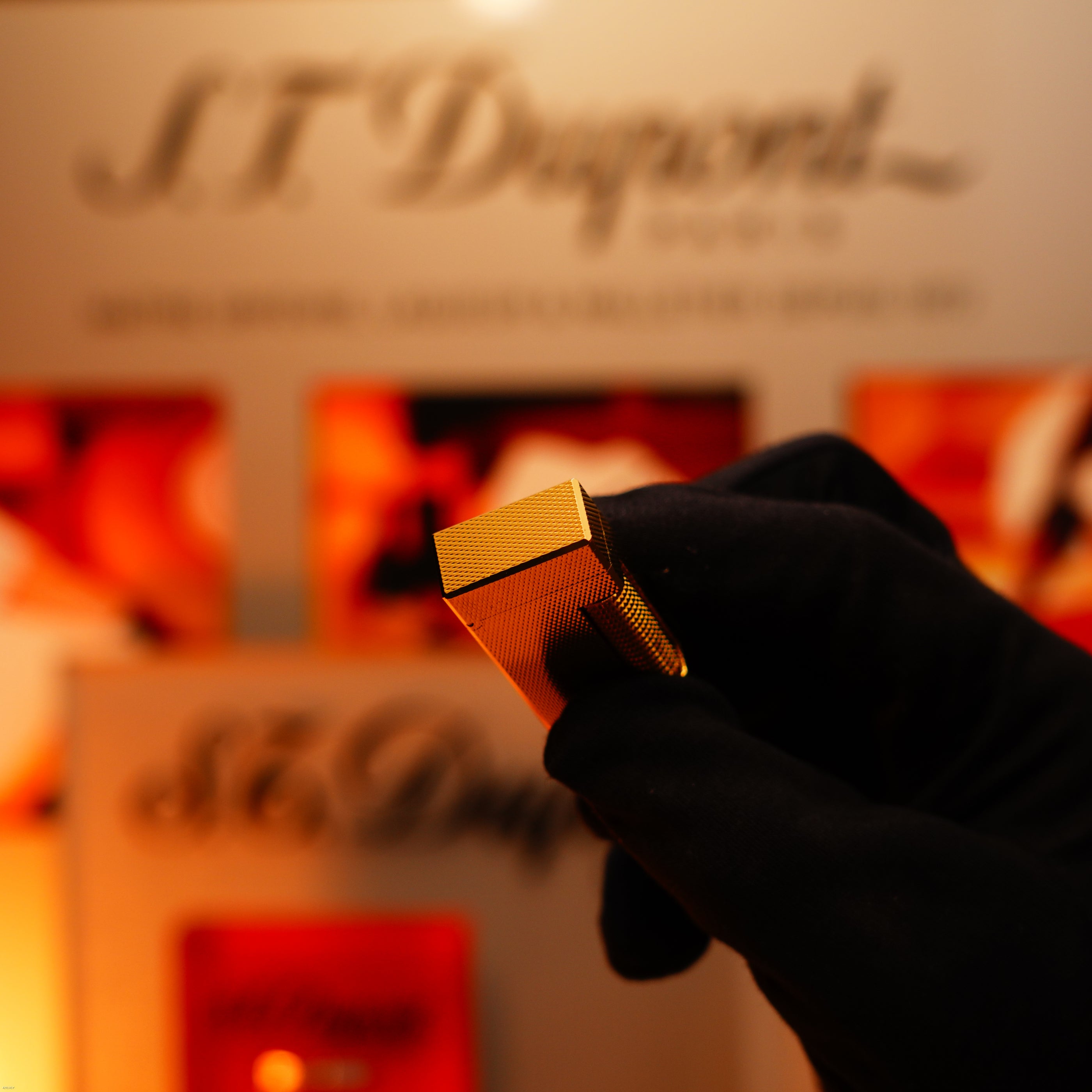 A gloved hand holds a Vintage 1975 Dunhill 24k Heavy Gold Finish Barley Pattern Early Version Lighter by Lucy Lighter before a blurred display featuring the brand's logo and signature branding, reflecting timeless elegance and quality.