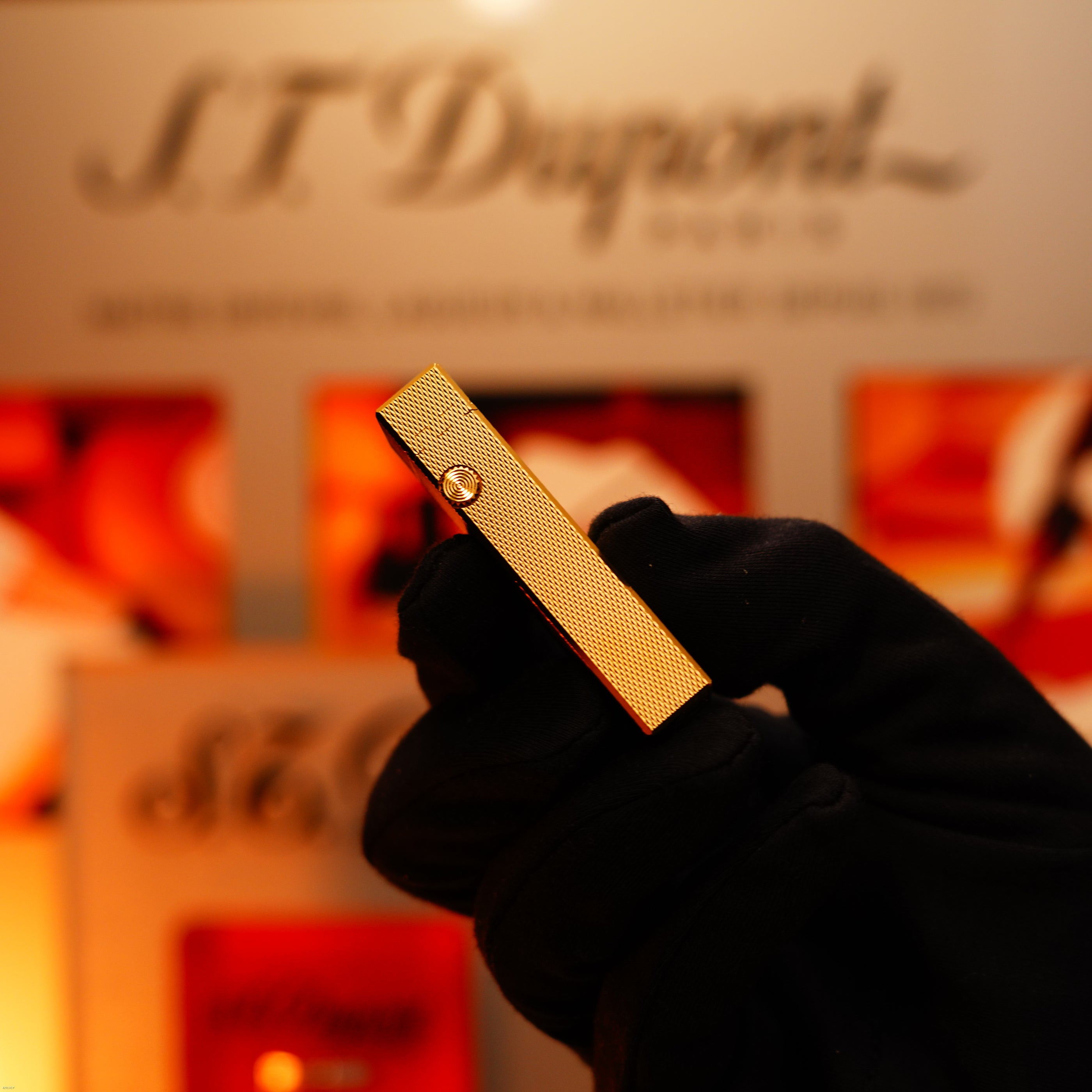 A gloved hand holds a vintage 1975 Dunhill 24k Heavy Gold Finish Barley Pattern Early Version Lighter by Lucy Lighter, showcased against a blurred background with the brand logo and display, emphasizing its quality guarantee and 12-month warranty.