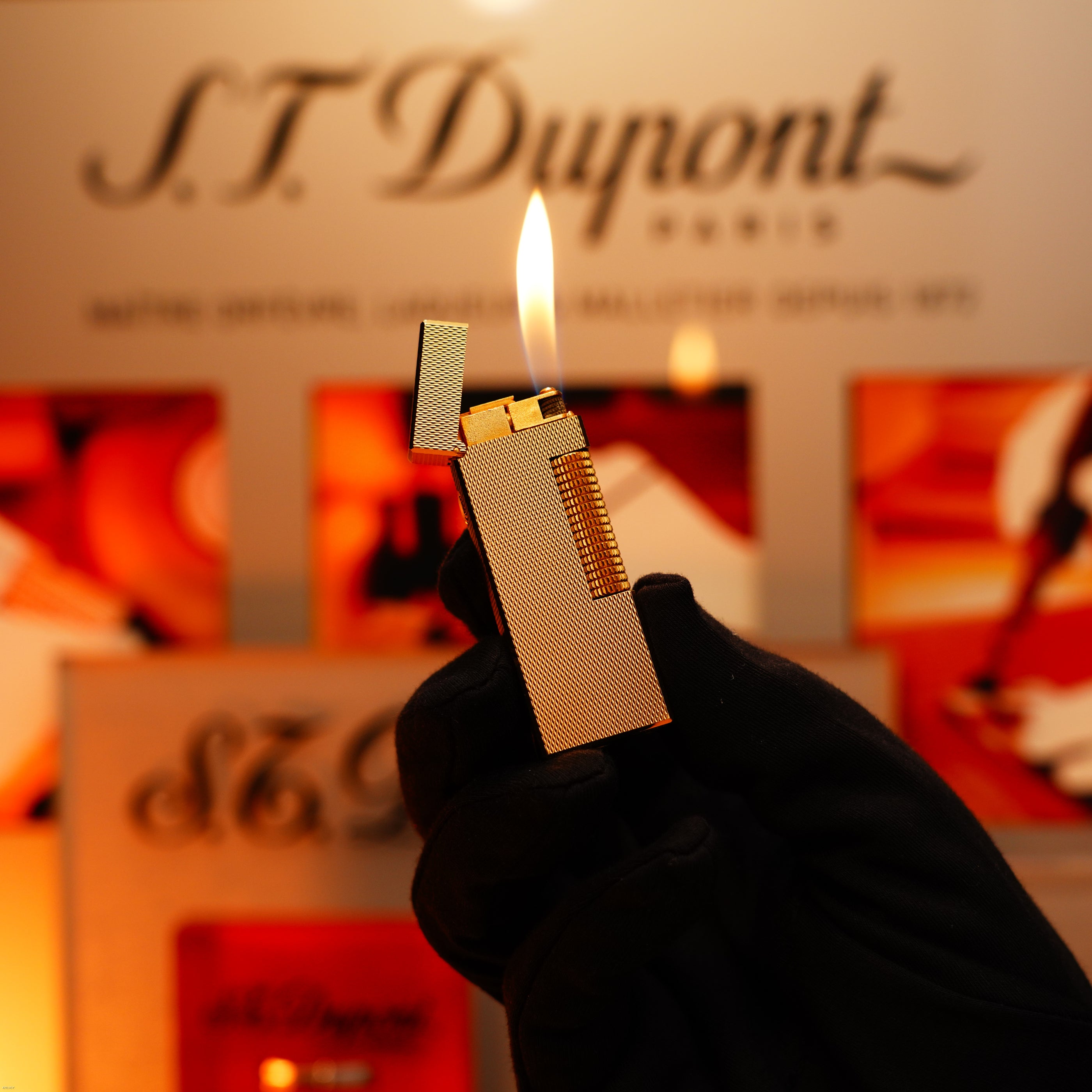 A gloved hand holds a Vintage 1975 Dunhill 24k Heavy Gold Finish Barley Pattern Early Version Lighter by Lucy Lighter, its lid open and flame lit, with a blurred Lucy Lighter display in the background—timeless style and quality.