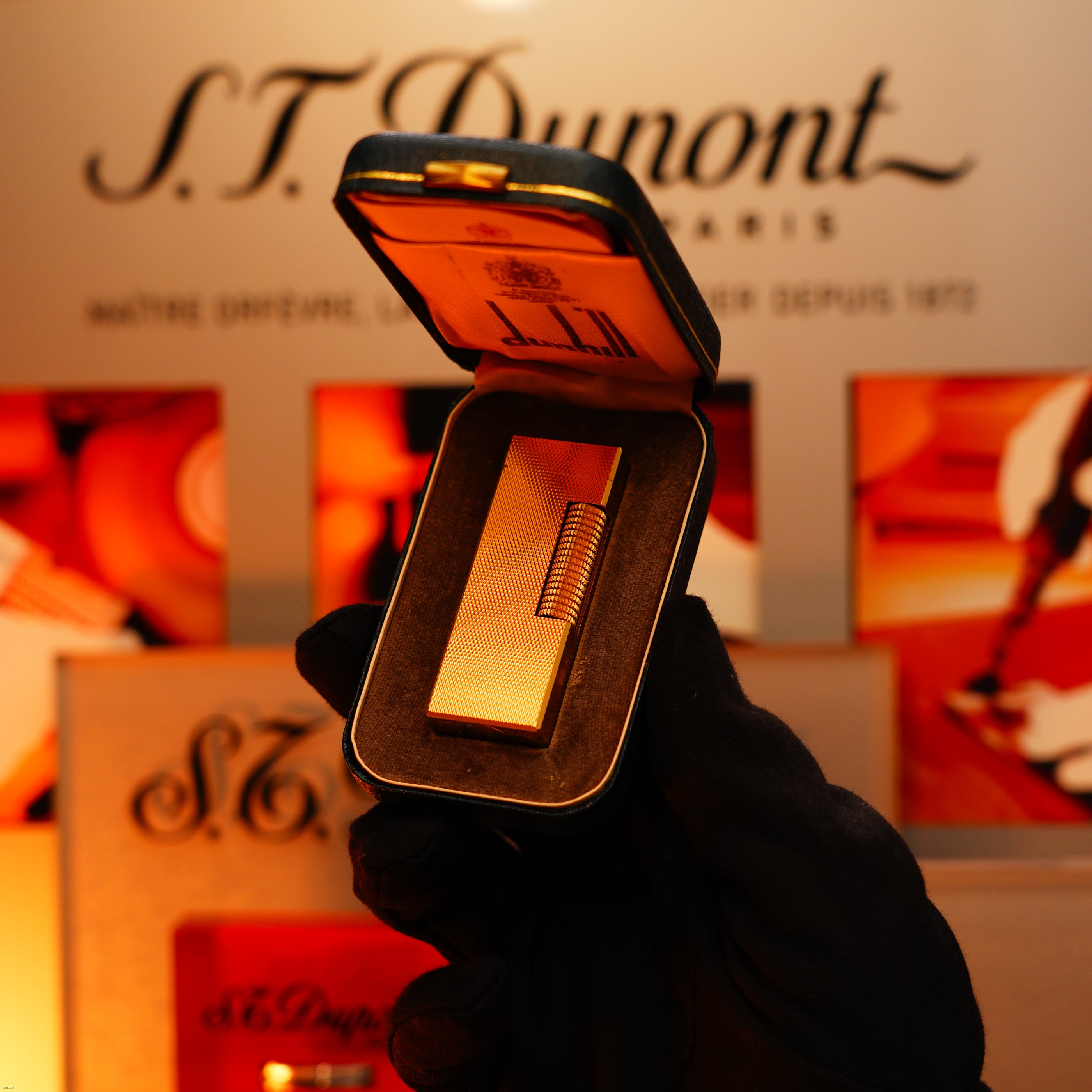 A gloved hand holds a Vintage 1975 Dunhill 24k Heavy Gold Finish Barley Pattern Early Version Lighter by Lucy Lighter in an open case, with the Lucy Lighter logo visible in the background.