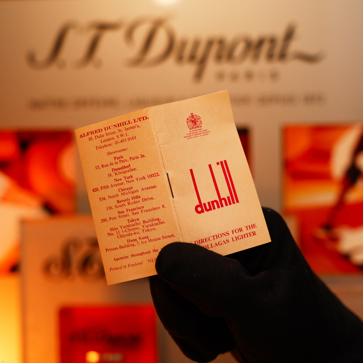 A gloved hand holds a Lucy Lighter card in front of an intage 1975 Dunhill 24k Heavy Gold Finish Barley Pattern Early Version Lighter, emphasizing its vintage luxury and collectible appeal.