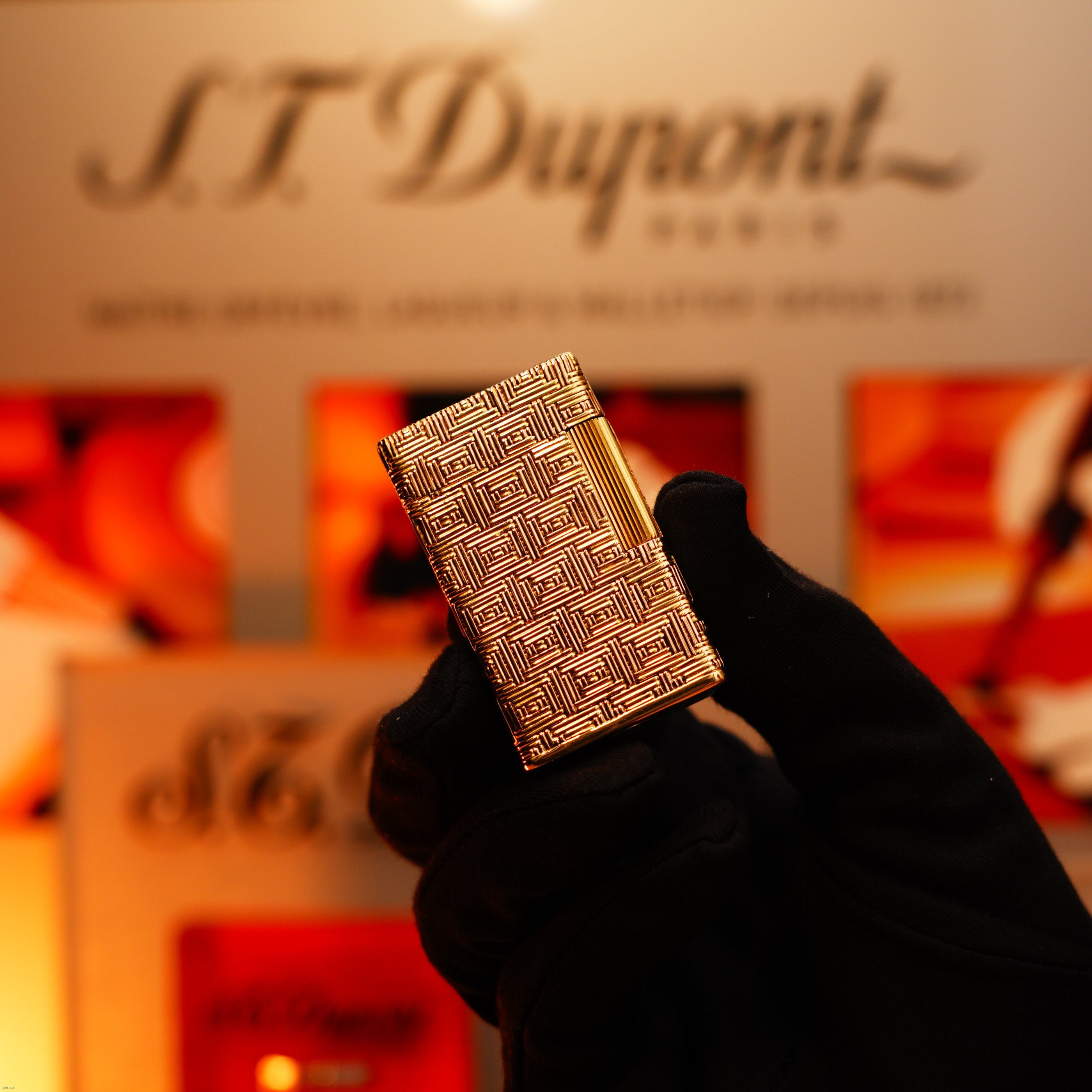 A gloved hand holds a solid 18K gold Vintage 1960 Extremely Rare 1/1 St Dupont Lighter by Lucy Lighter, heavy early edition with 750 hallmark, against a blurred branded display background.