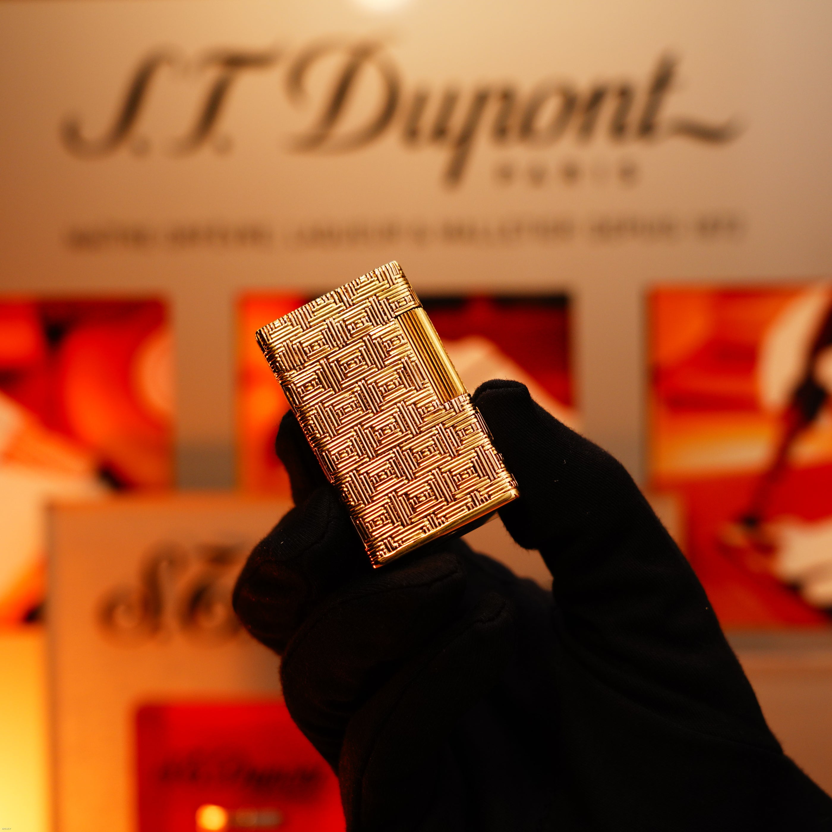 A gloved hand holds a Vintage 1960 Extremely Rare 1/1 St Dupont Lighter by Lucy Lighter—solid 18K gold, heavy early edition with 750 hallmark—showcasing exclusive elegance before a blurred display.