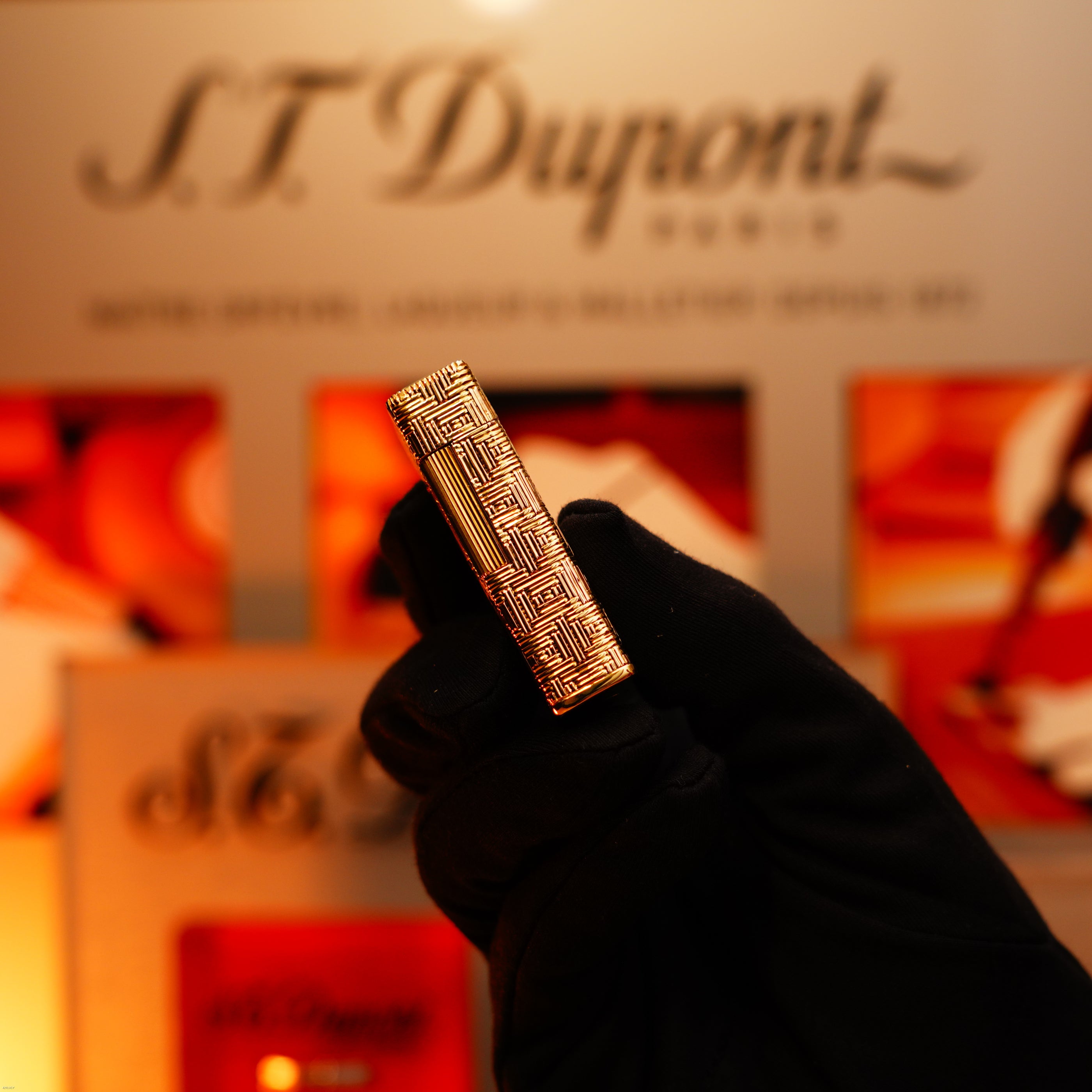 A gloved hand holds the Lucy Lighter Vintage 1960 Extremely Rare 1/1 St Dupont Lighter, crafted in solid 18K gold and heavy early edition with 750 hallmark, in front of a blurred display.