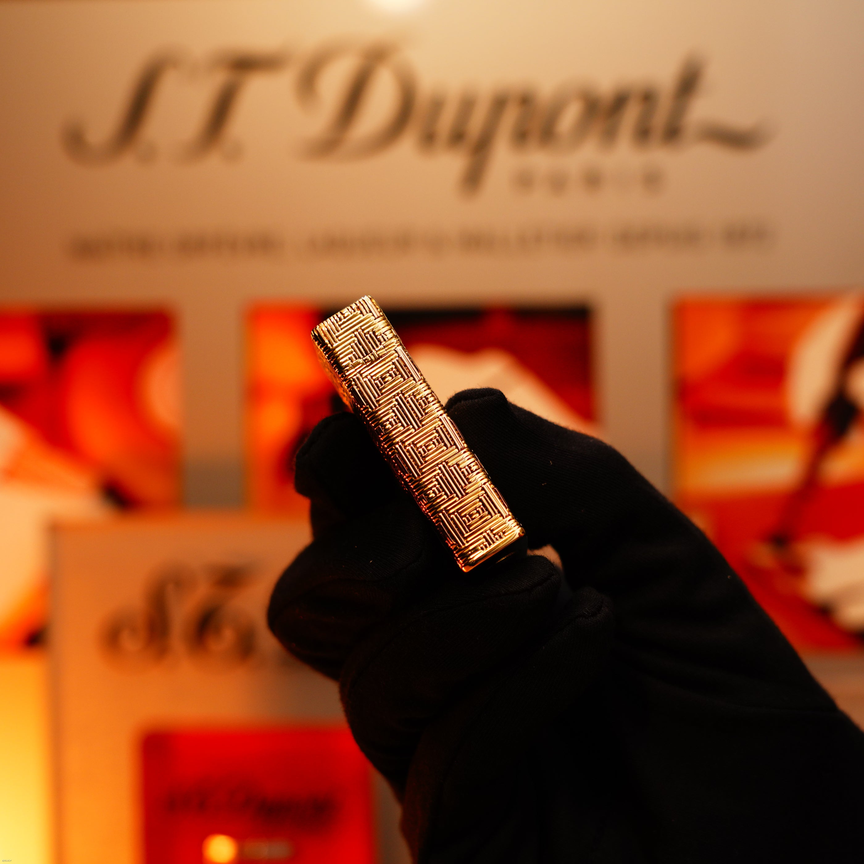 A gloved hand holds a Vintage 1960 Extremely Rare 1/1 Lucy Lighter St Dupont solid 18K gold lighter, heavy early edition with 750 hallmark, geometric pattern, branded packaging beside it and a blurred background.
