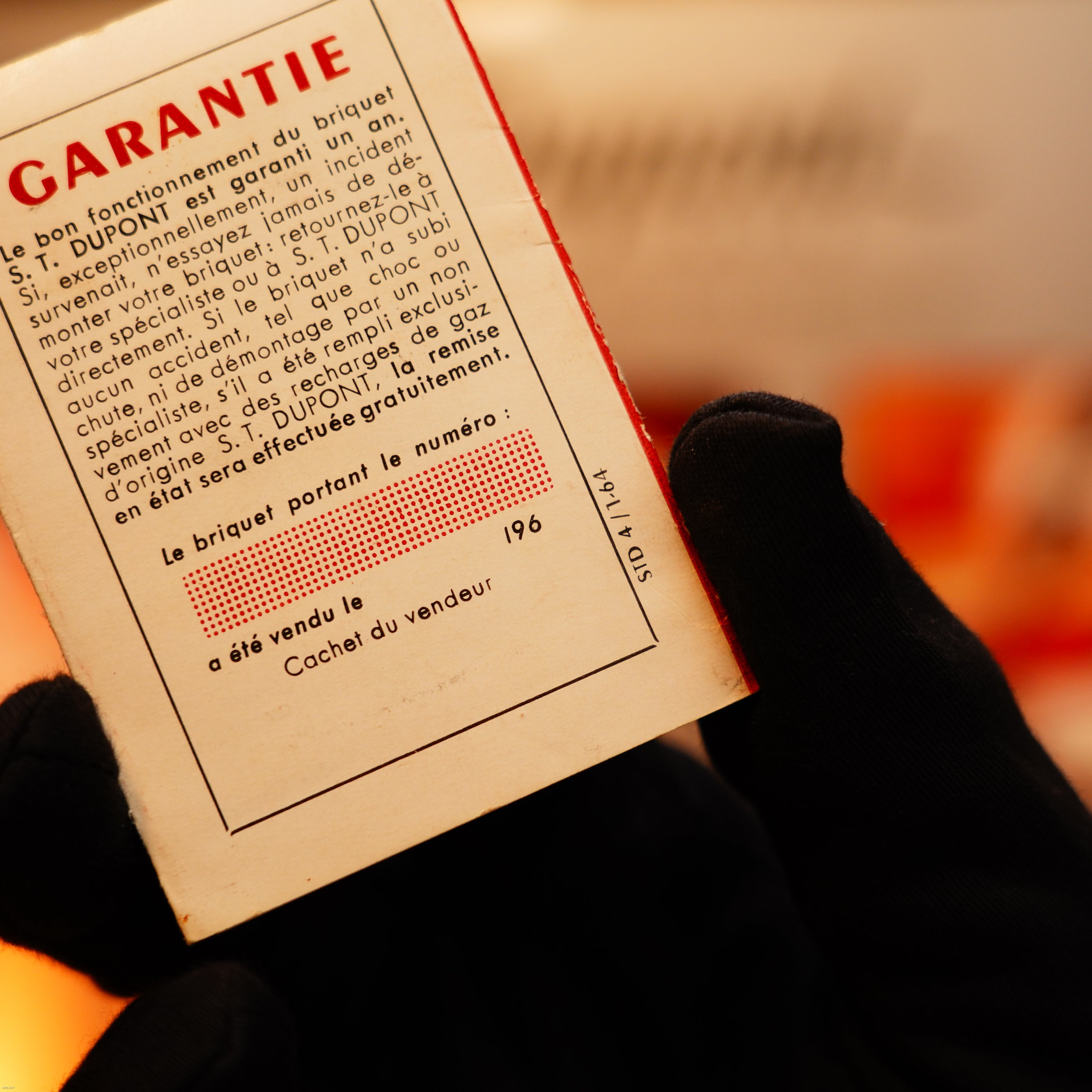 A gloved hand holds a French warranty card for the Vintage 1960 Extremely Rare 1/1 St Dupont Lighter by Lucy Lighter—solid 18K gold, heavy early edition with 750 hallmark—against an orange blurred background.