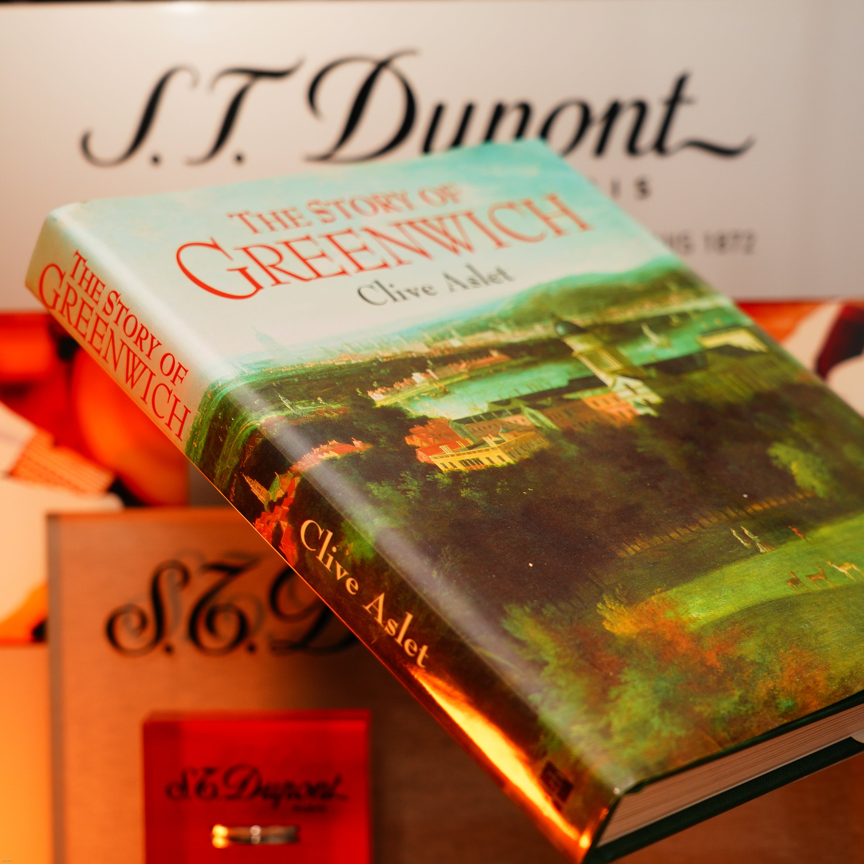 A hardback copy of "The Story of Greenwich" by Clive Aslet rests on a surface, with Lucy Lighter branding and a red box—suggesting the Vintage Extremely Rare Alfred Dunhill Full Set GMT Solid Stainless Steel lighter—in the background.