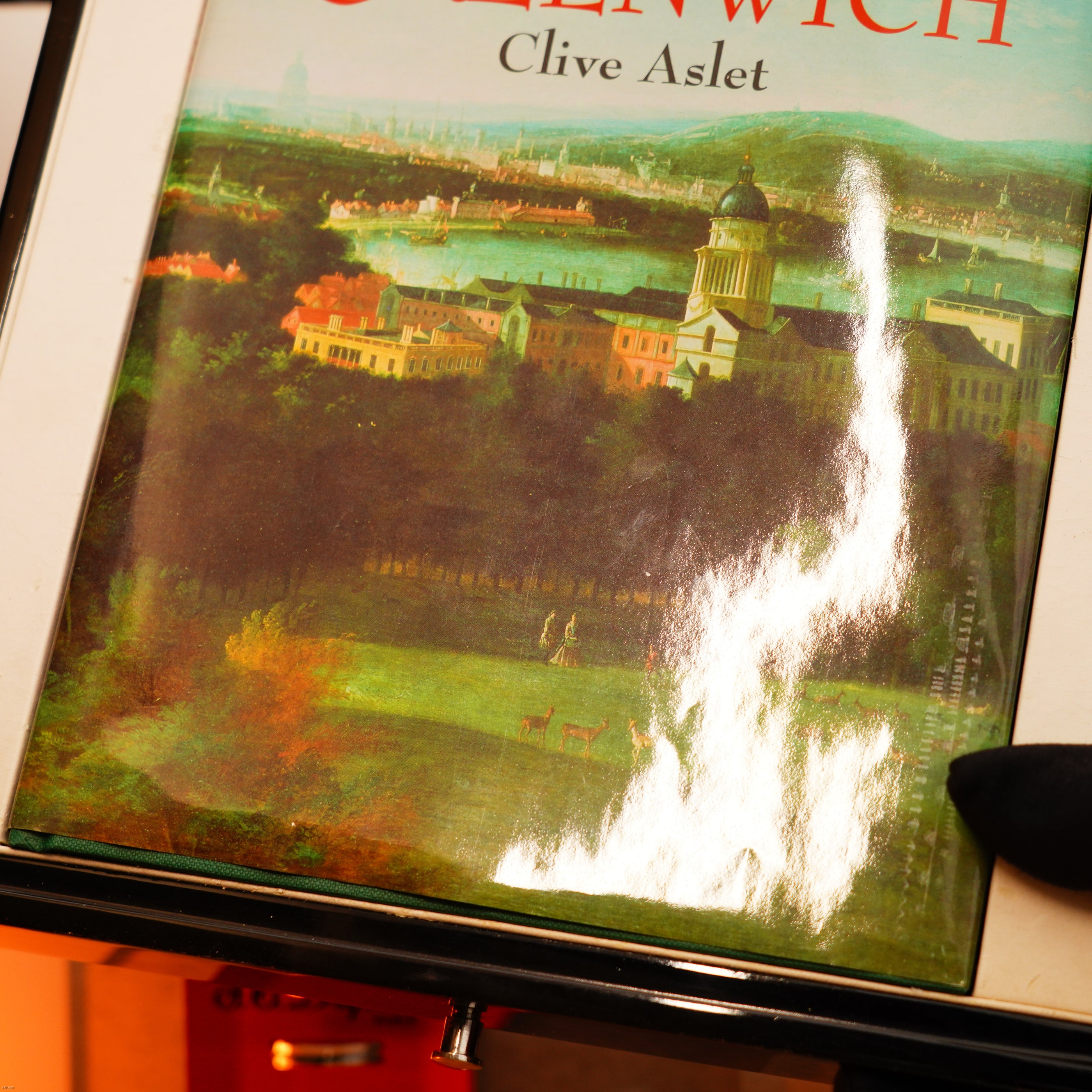 A book titled "Greenwich" by Clive Aslet is displayed, its cover showing Greenwich scenes. Next to it is a Vintage Extremely Rare Alfred Dunhill Full Set GMT Solid Stainless Steel lighter by Lucy Lighter, near a gloved hand.