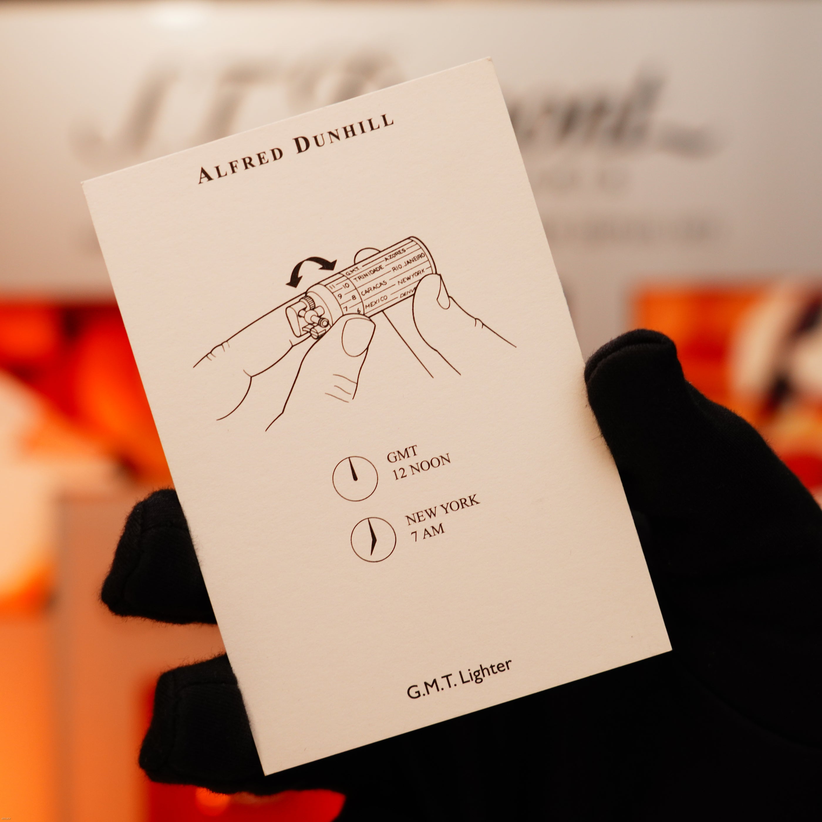 A gloved hand holds a Vintage Extremely Rare Alfred Dunhill Full Set GMT Full Solid Stainless Steel card by Lucy Lighter, showing a vintage lighter diagram and clocks marked GMT 12 Noon and New York 7 AM, labeled “G.M.T. Lighter.”.