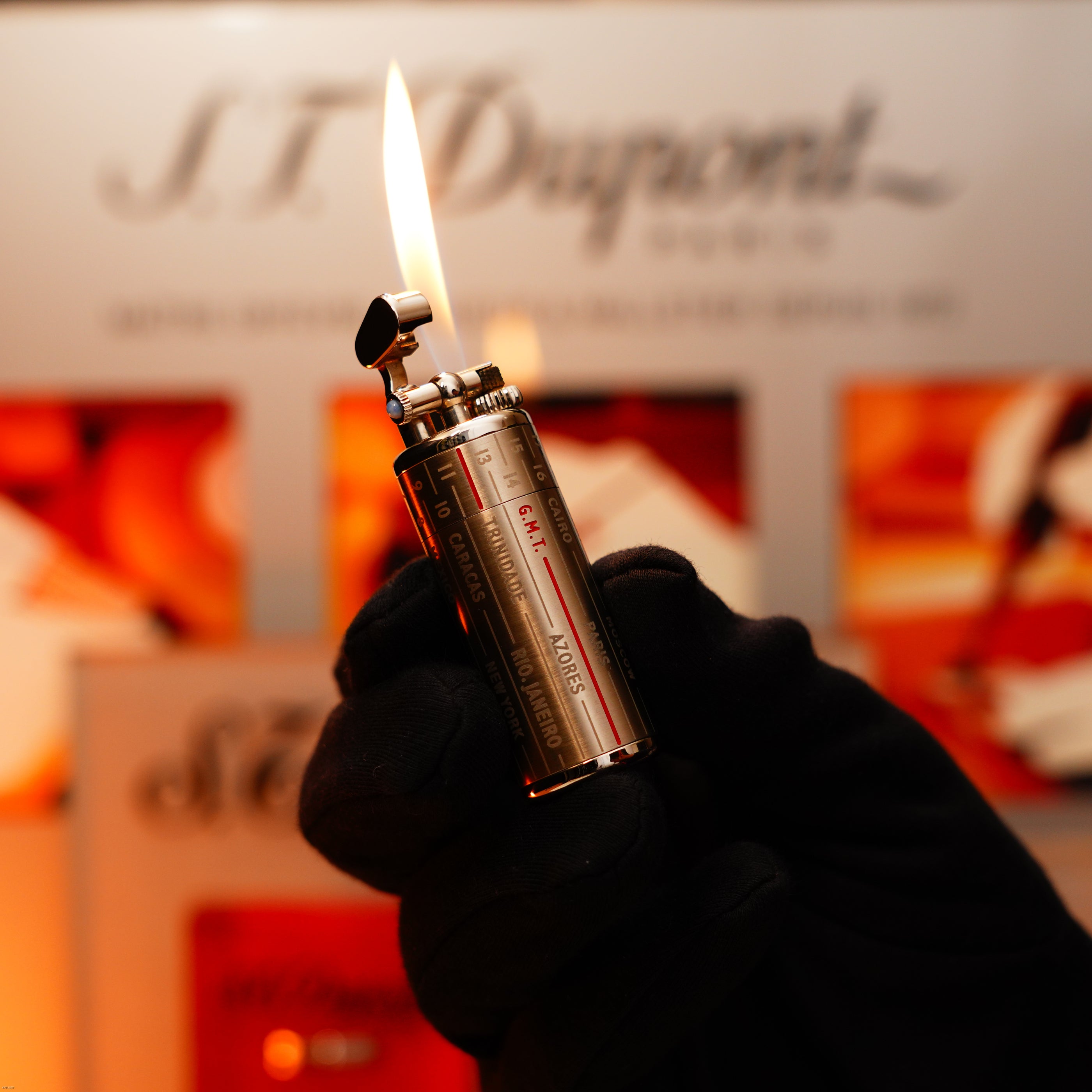 A gloved hand holds a lit Lucy Lighter Vintage Extremely Rare Alfred Dunhill Full Set GMT Solid Stainless Steel with Alfred Dunhill Signature; the flame is visible against a blurred background with branded signage.